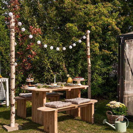 Ein gedeckter Gartentisch  Emilia mit den passenden Bänken steht unter der Birkenstamm Lichterkette in einem sommerlichen Garten.