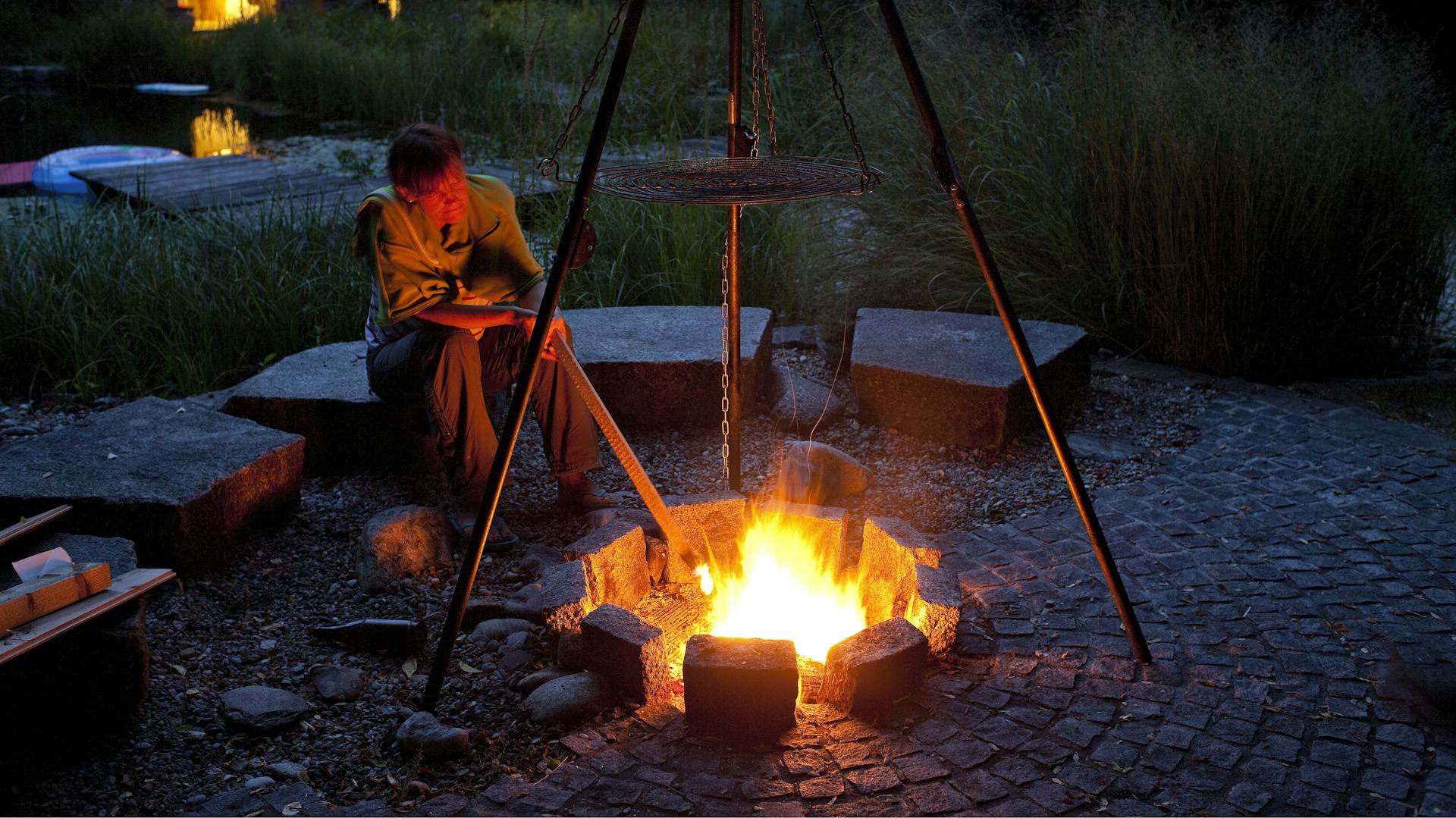 Person sitzt abends an Feuerstelle mit Lagerfeuer, Dreibein und Sitzgelegenheiten