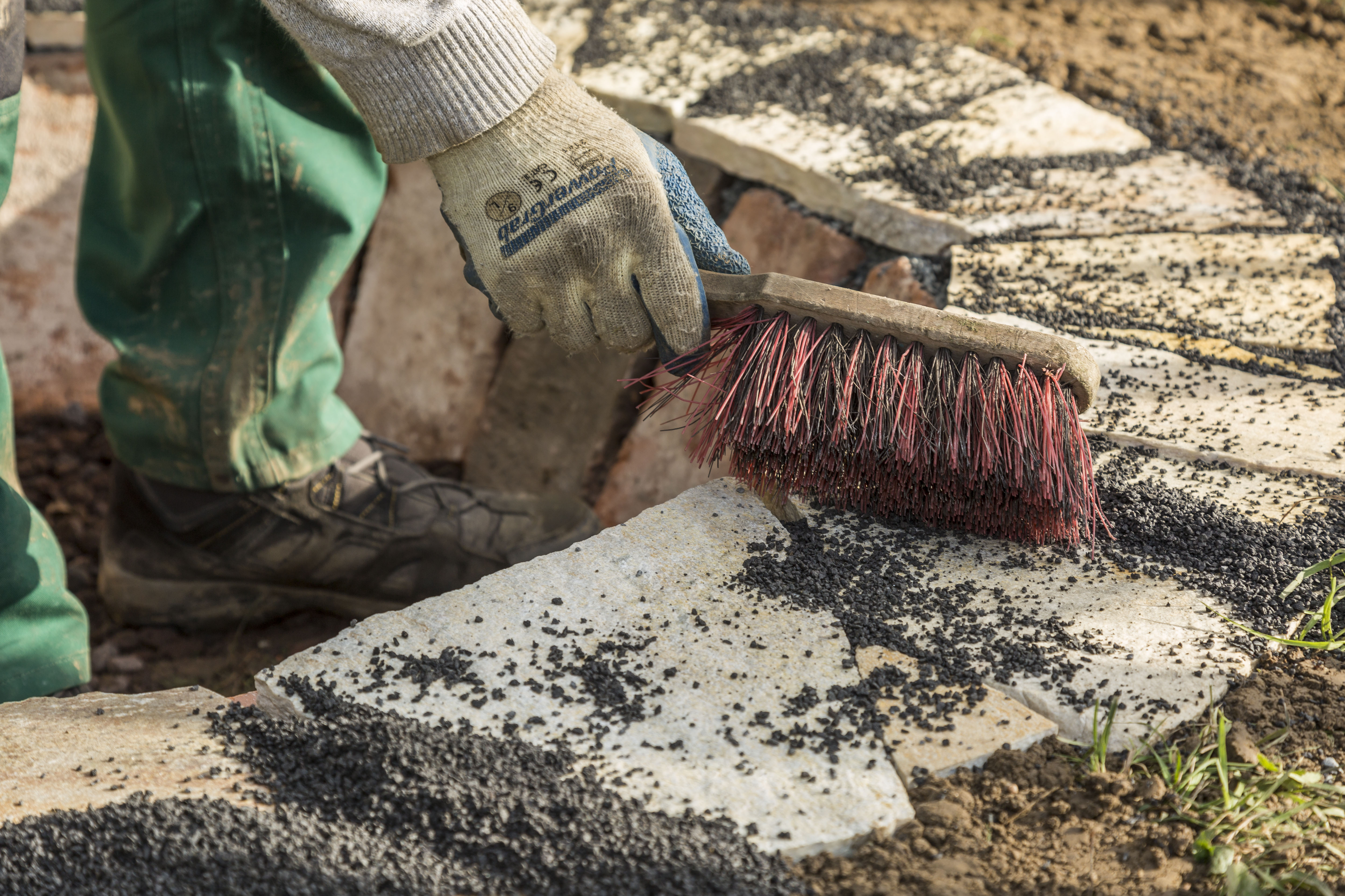 Osoba špáruje medzery medzi obrubníkmi pomocou ručnej kefy s posypom.
