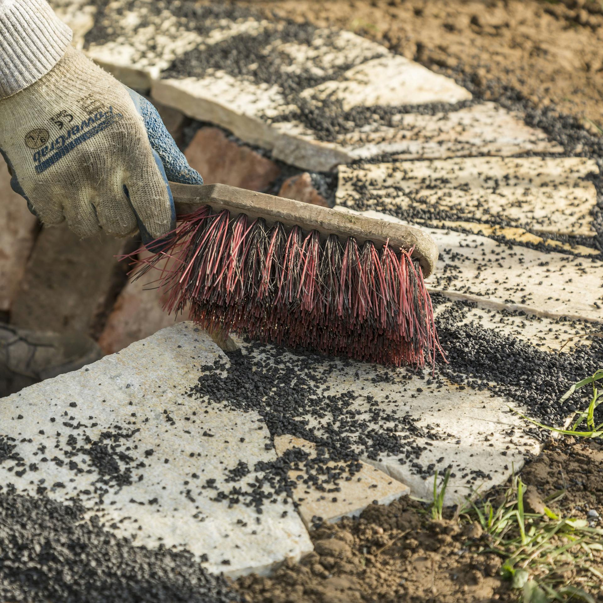 Person verfugt die Lücken zwischen den Randsteinen mittels eines Handfegers mit Splitt.