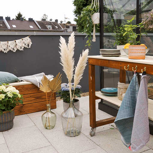 Bank Alfred und Servierwagen Sophie stehen auf einer mit Vasen und Trockenblumen geschmückten Dachterrasse.