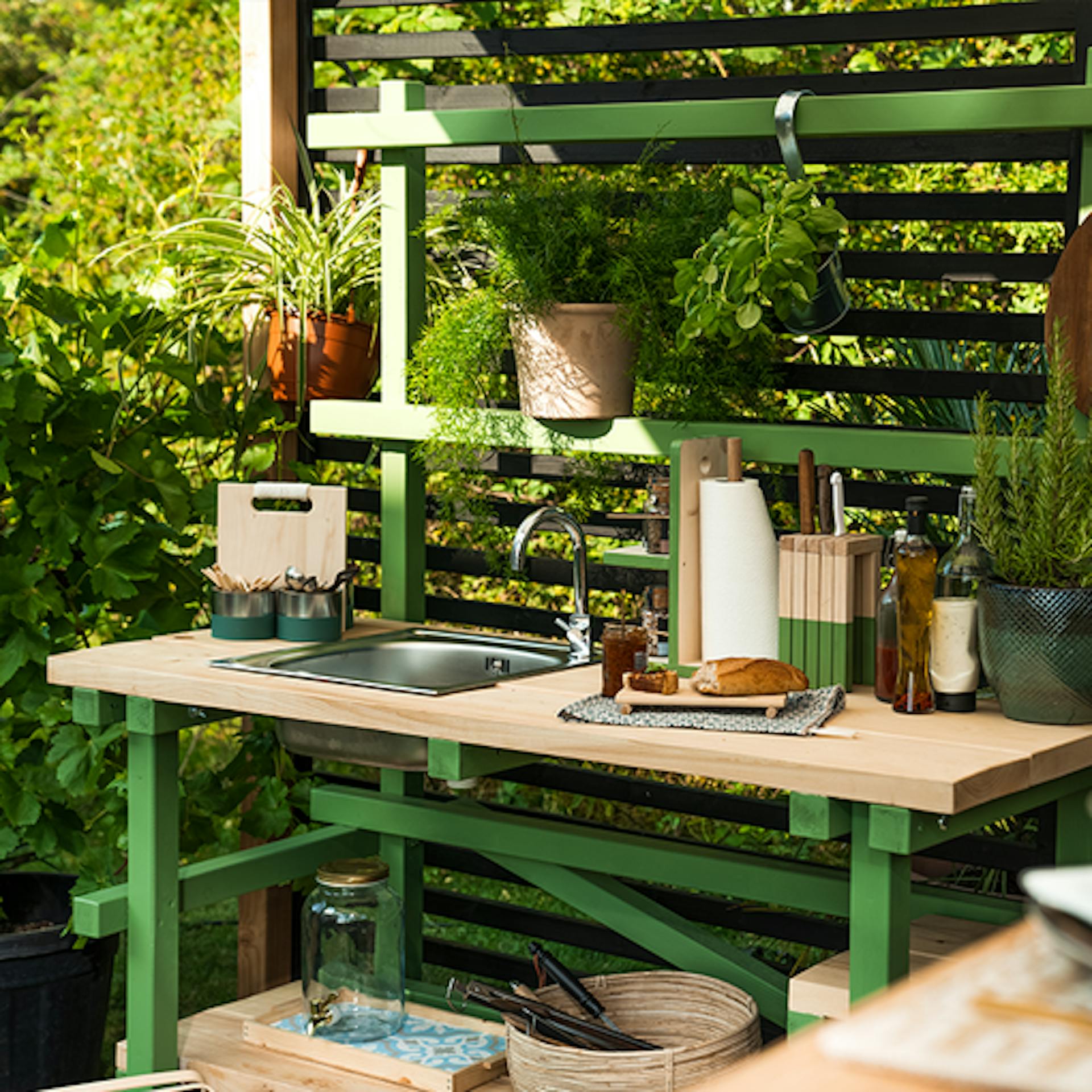 Outdoorküche Alfons, welche hellgrün gestrichen worden ist, steht in einer Ecke eines sommerlichen Gartens.