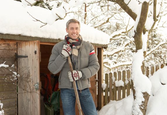Uomo in giacca invernale davanti a una capanna di legno coperta di neve con una pala da neve in mano.