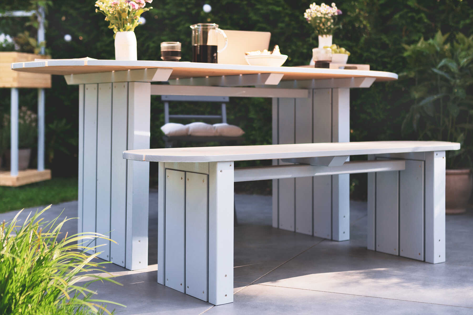 Gartenbank Emilia steht auf der Gartenterrasse und ist mit Blumen und Kissen dekoriert.