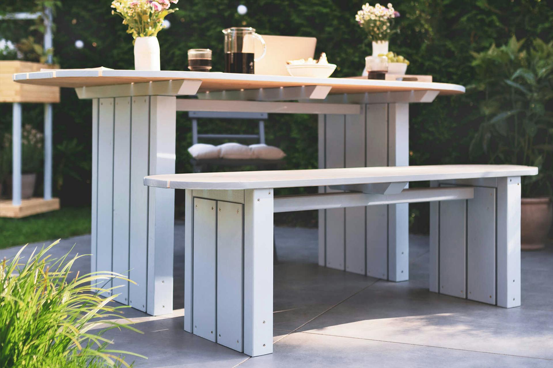 Gartenbank Emilia steht auf der Gartenterrasse und ist mit Blumen und Kissen dekoriert.