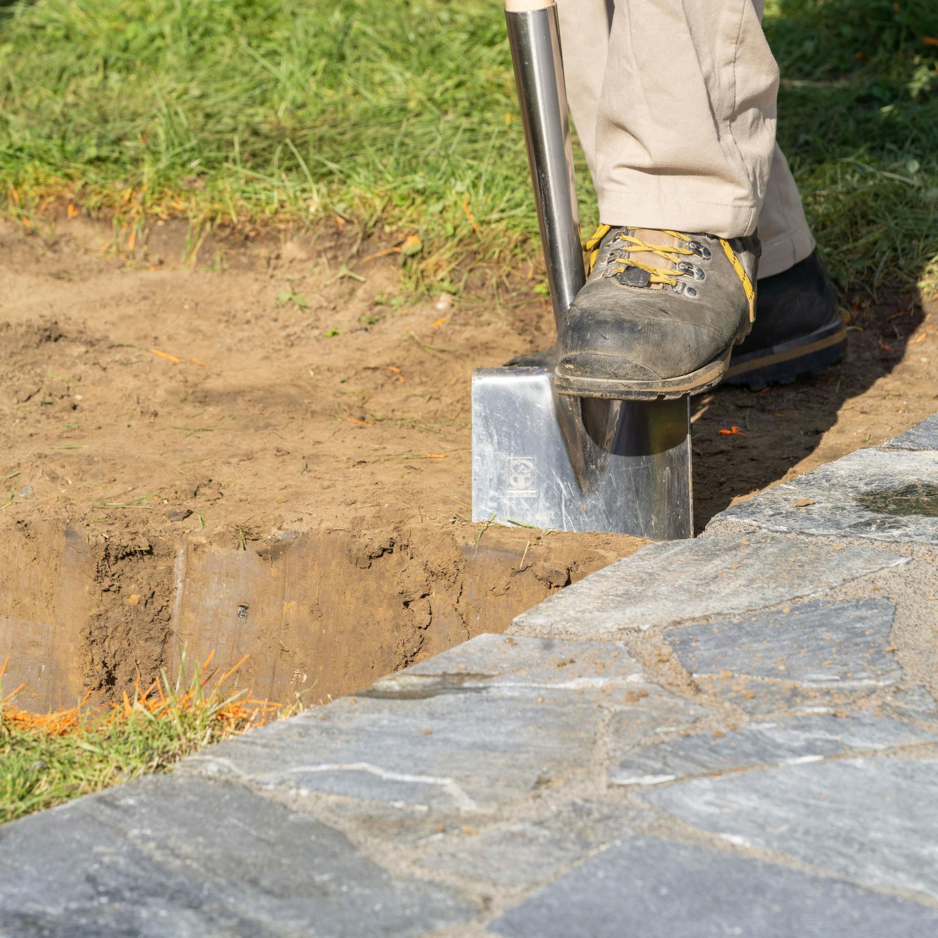Mann steht mit dem Fuß auf einem Spaten, der in der Erde steckt