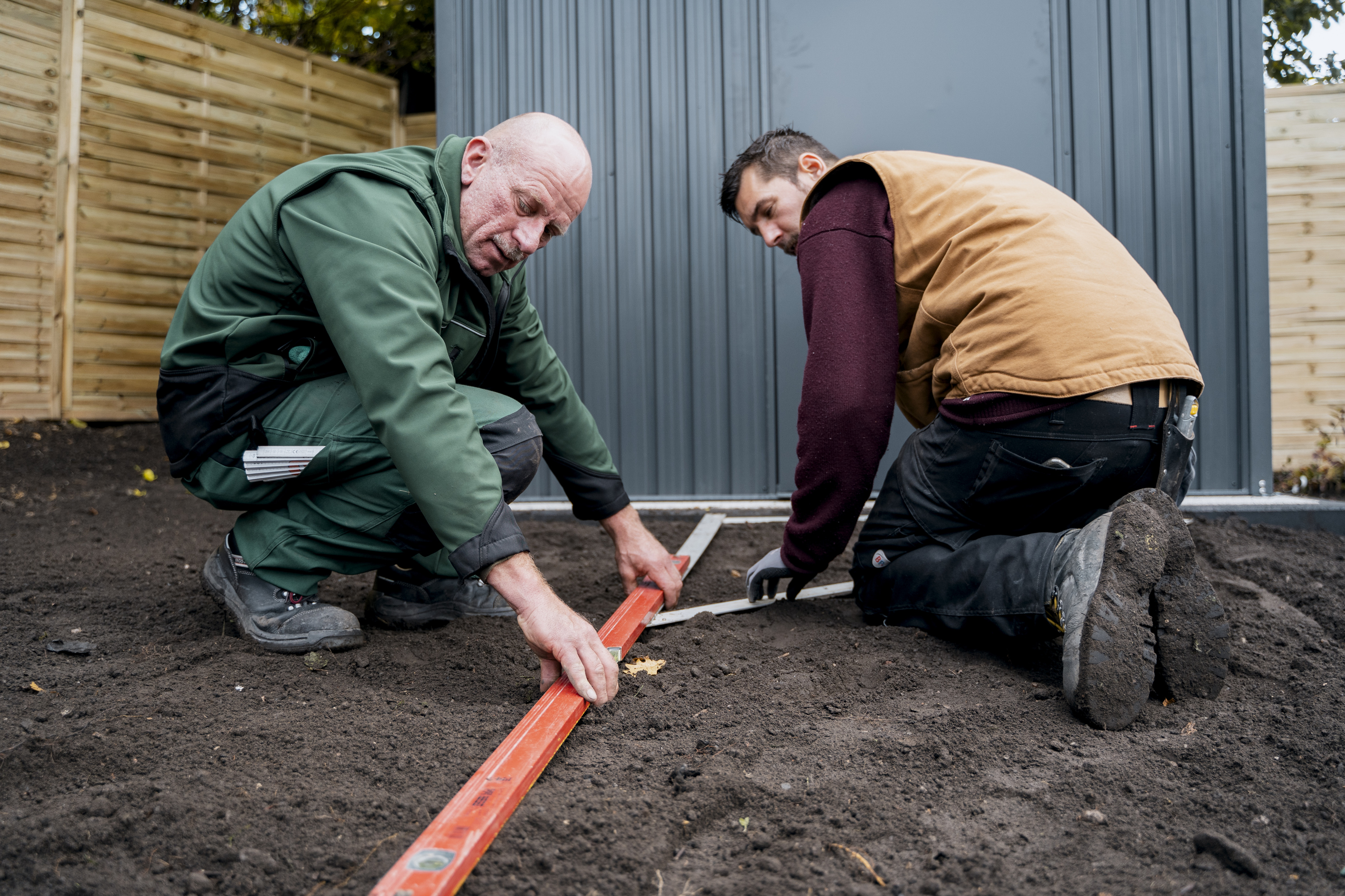 Két férfi szögmérővel és mérőléccel méri a kavicsos út helyét.