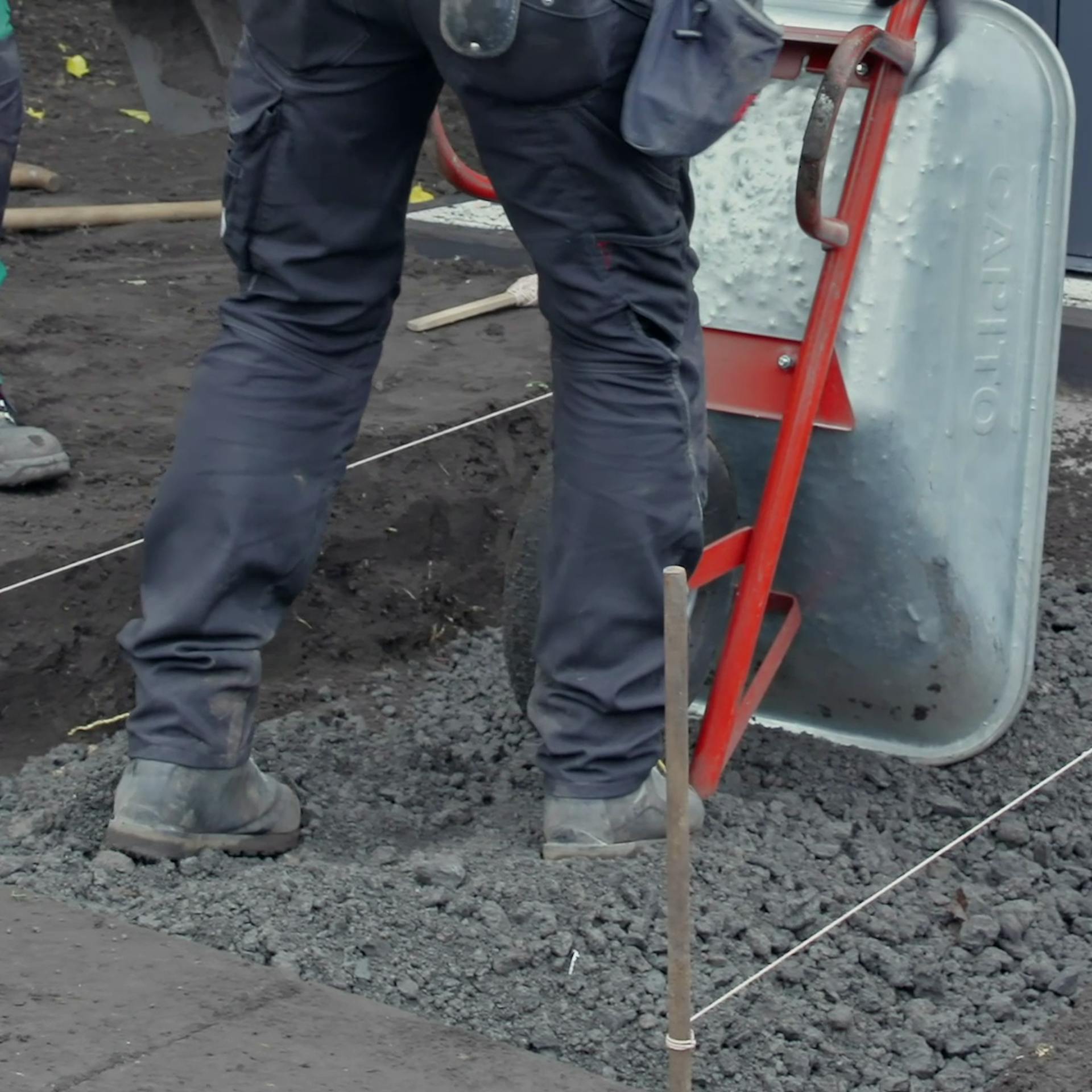 Person schüttet Schotter mit Scheibtruhe in ausgehobene Grube für Gartenweg.
