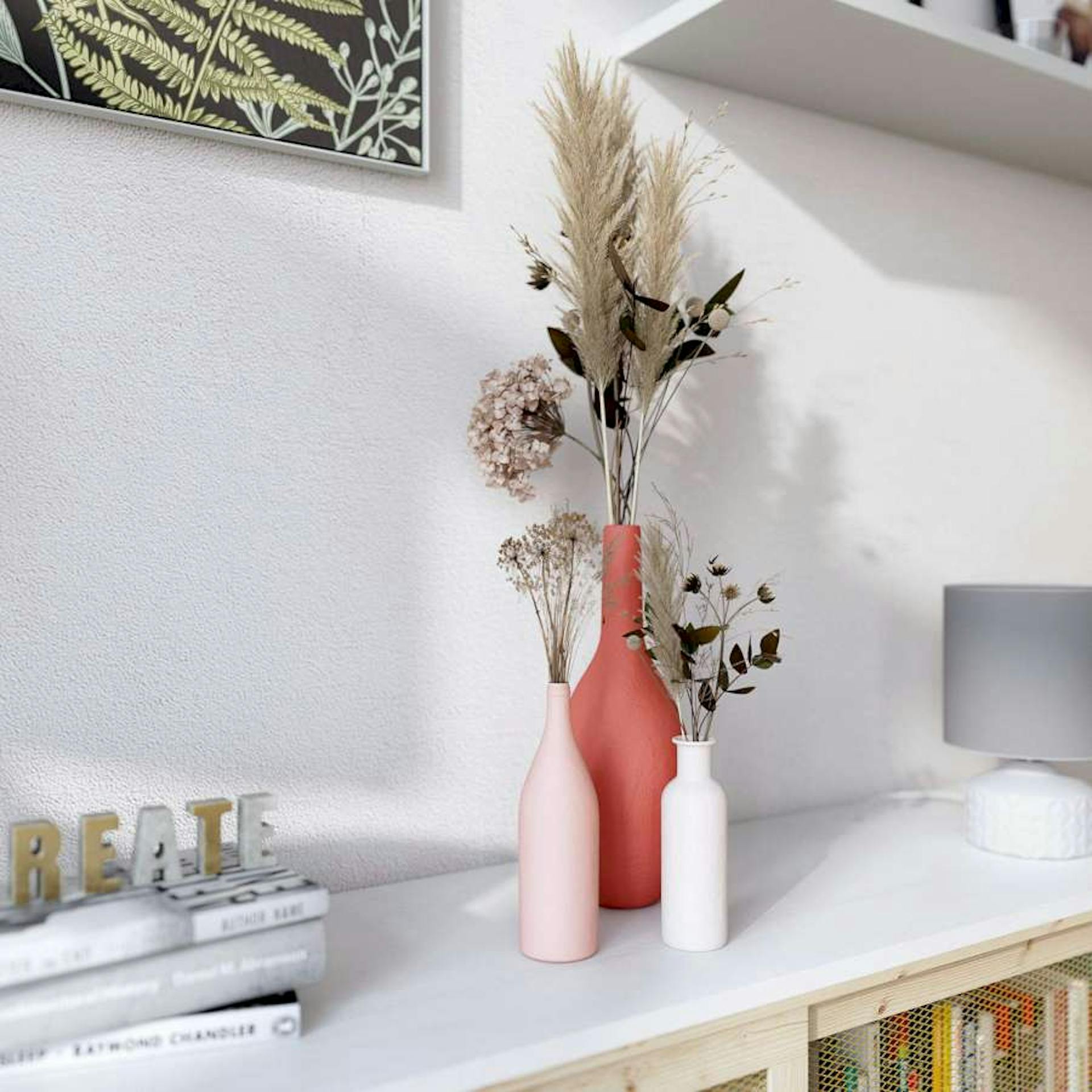 Auf einem weißen Regal stehen Vasen mit Trockenblumen im Faux Ceramic Stil. Daneben befinden sich Bücher und eine Lampe.