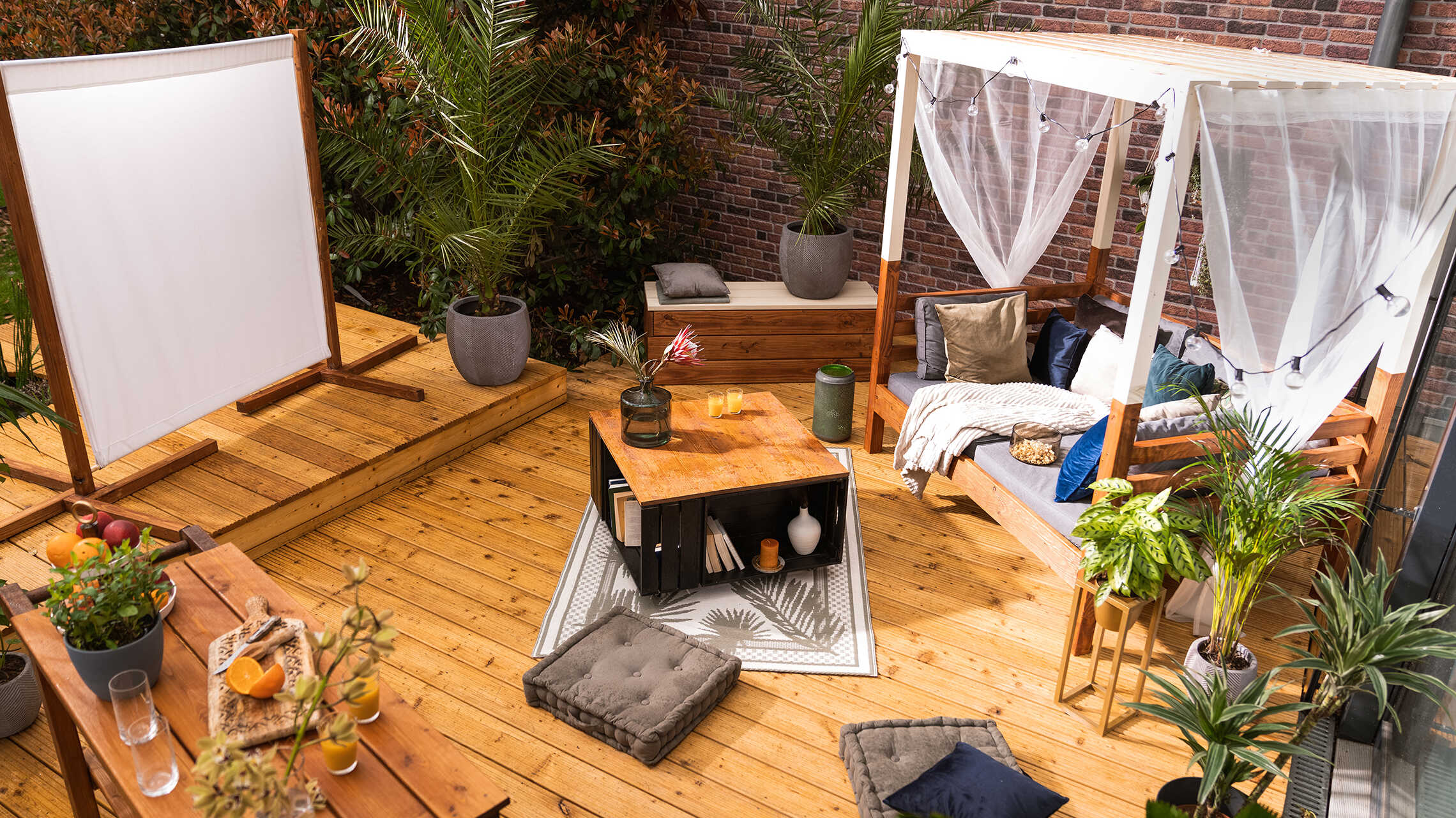 Eine Terrasse auf der eine Leinwand vor die Sonneninsel Helena platziert wurde und zwischen den beiden eine Bank Alfred und ein Couchtisch Vierfach stehen.