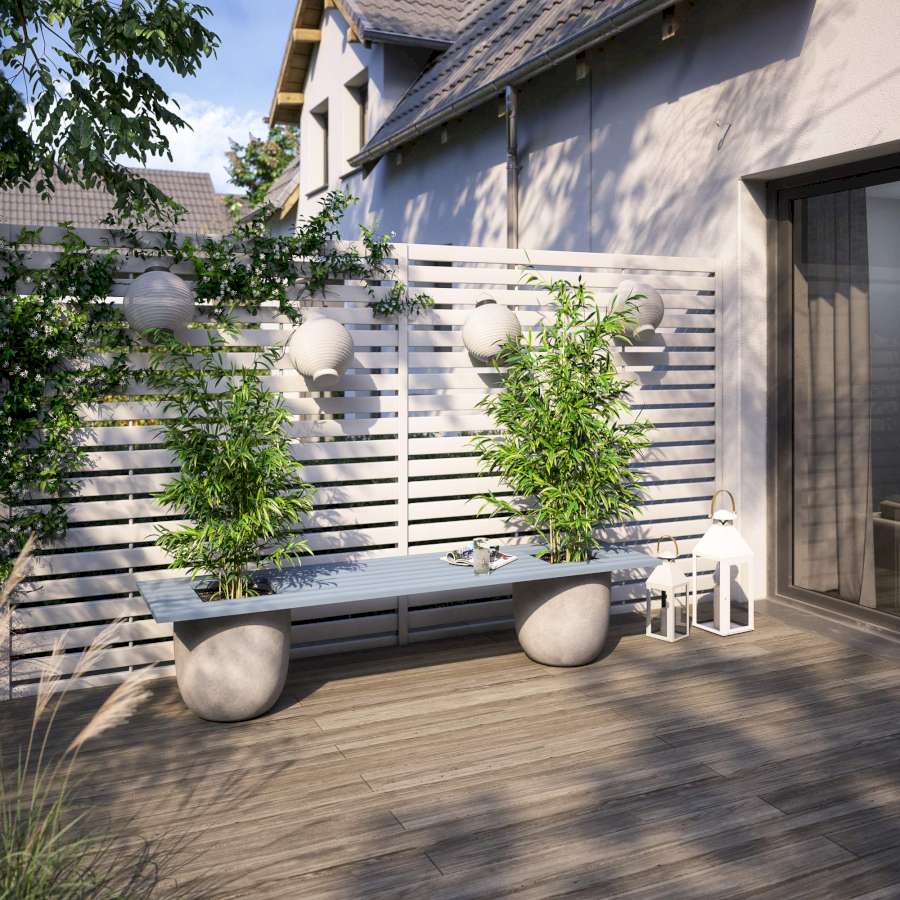 Eine Gartenbank, die als Beine Pflanzkübel verwendet, steht vor einem weißen Zaun auf einer Terrasse.