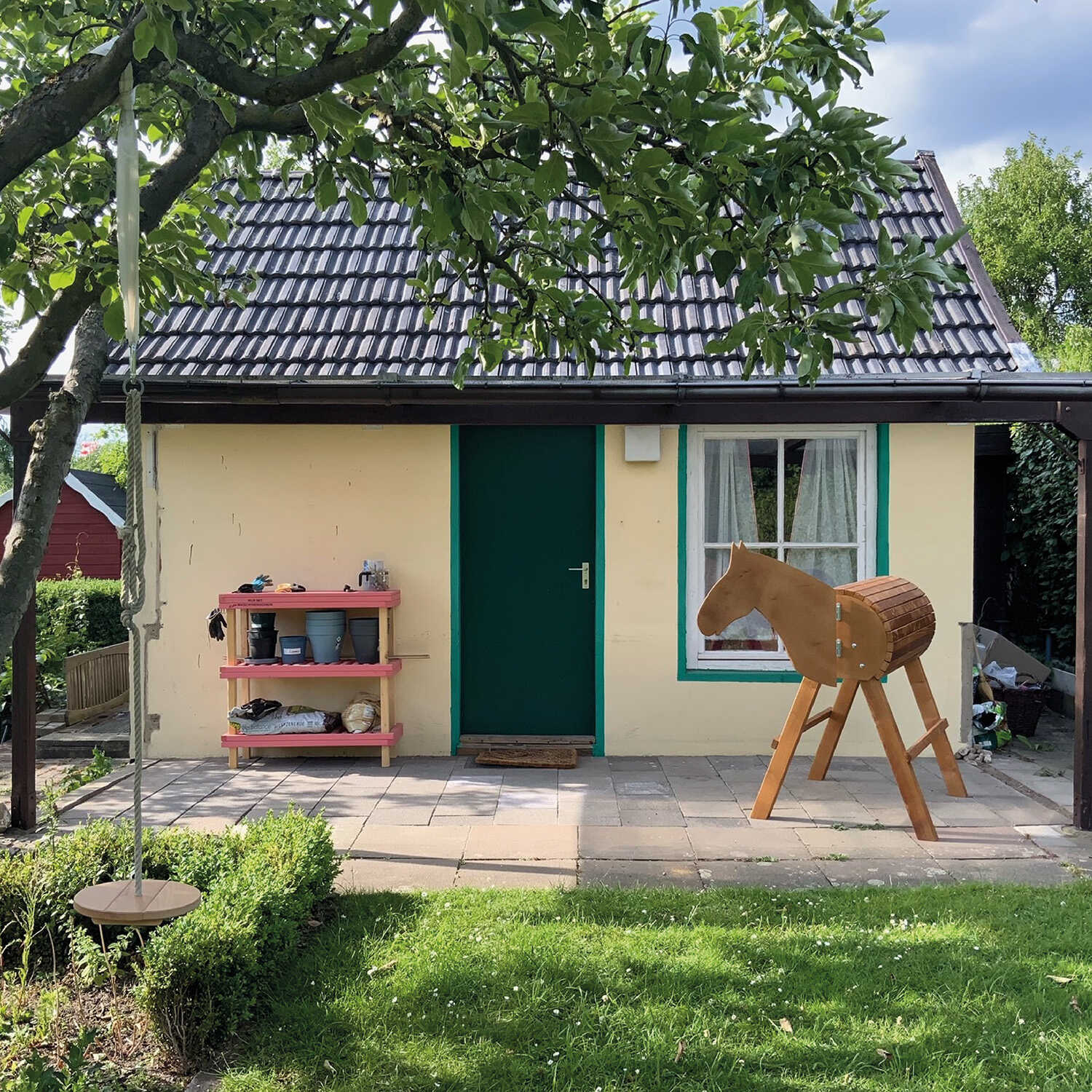 Vor einem kleinen, gelben Häuschen mit grünen Akzenten steht ein Regal mit Blumentöpfen und ein Holzpferd.