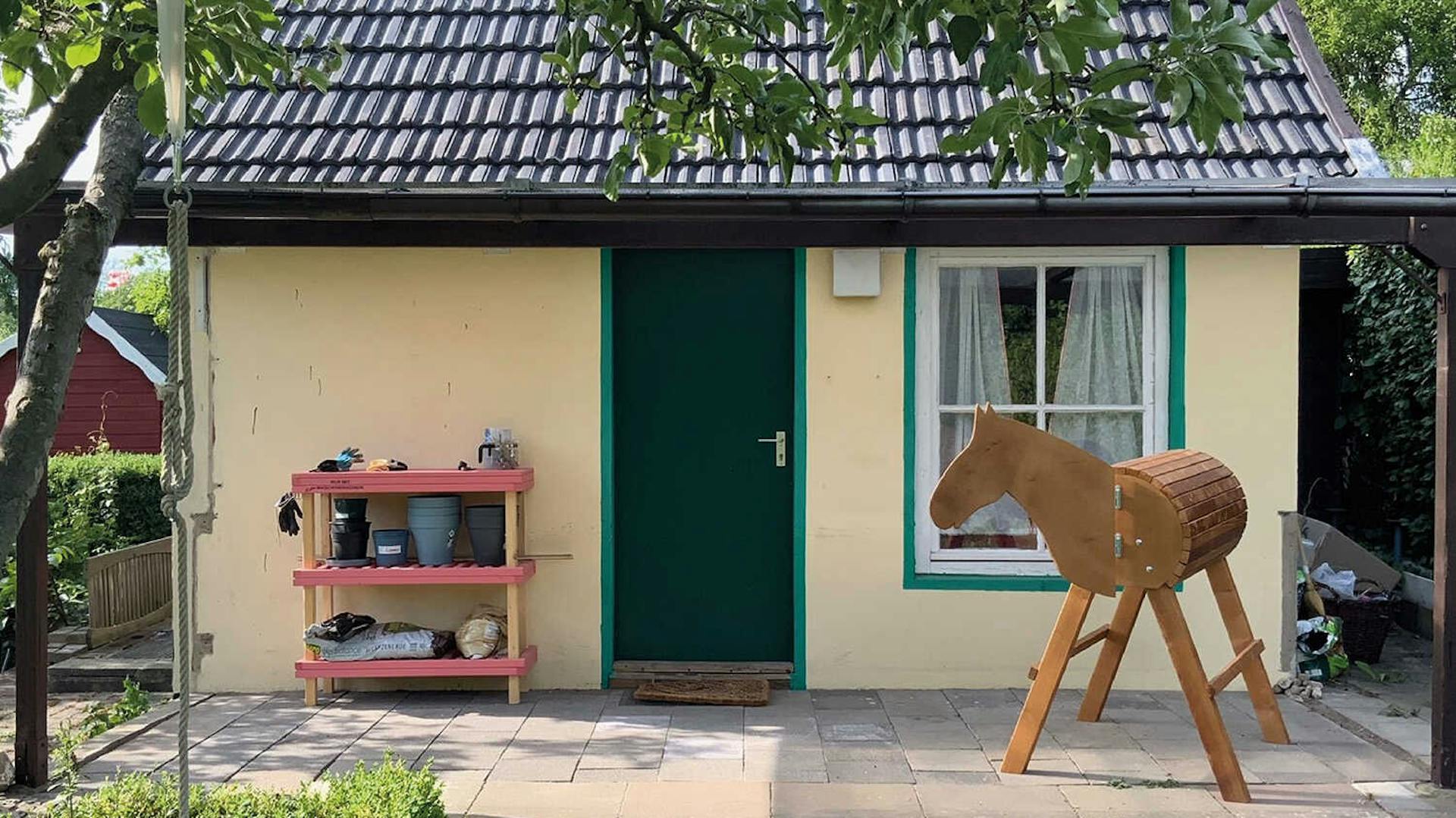 Vor einem kleinen, gelben Häuschen mit grünen Akzenten steht ein Regal mit Blumentöpfen und ein Holzpferd.