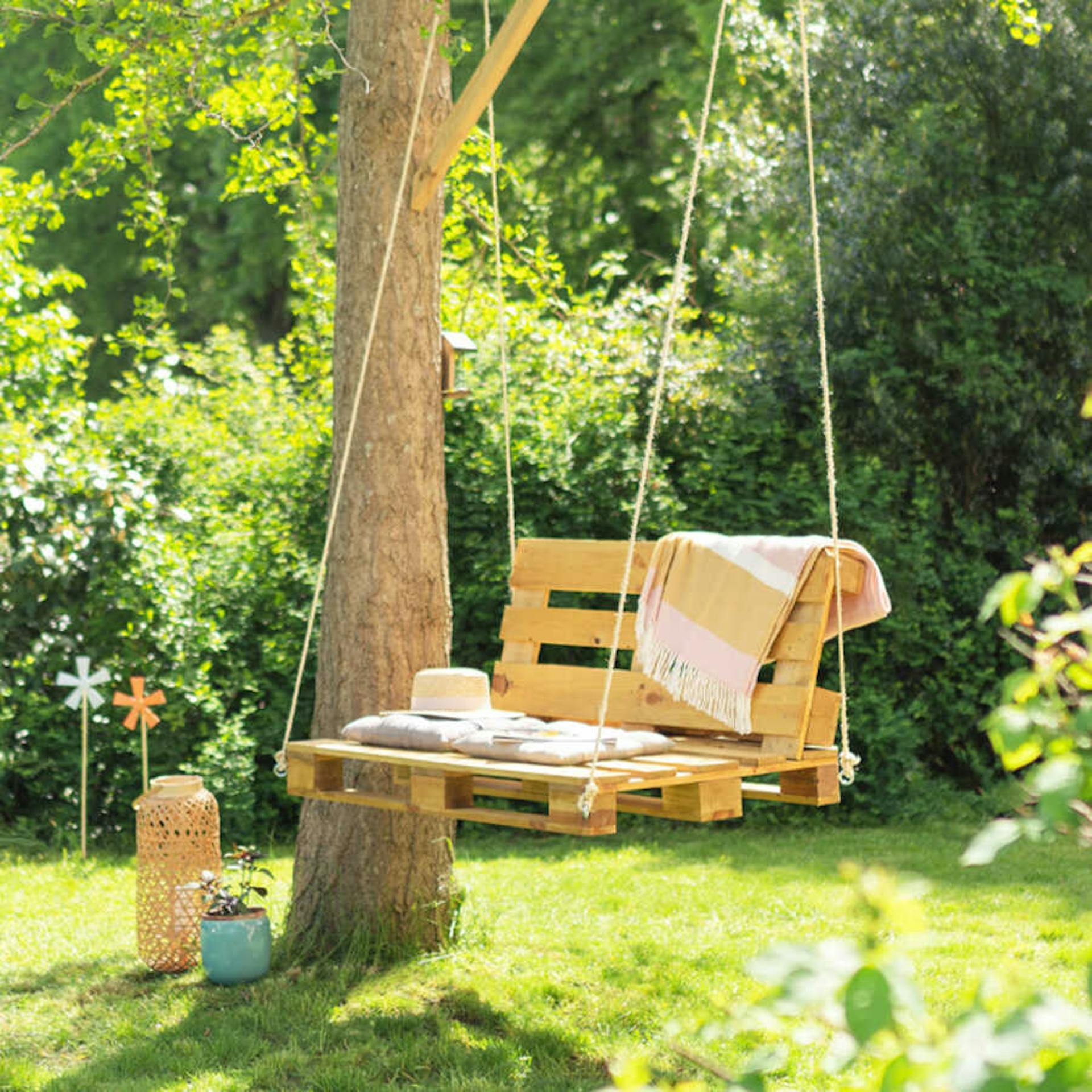 Die Palettenschaukel Baumelig hängt in einem Garten an einem Baum, neben dem ein Blumentopf und ein Windlicht stehen.