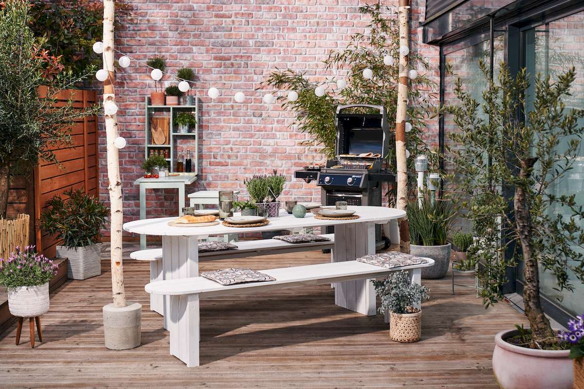 Eine Lichterkette ist an zwei Birkenstämmen befestigt wurden, diese stehen auf einer Terrasse mit um einem gedeckten Tisch. Um den Tisch herum befinden sich viele Pflanzen und ein Grill.