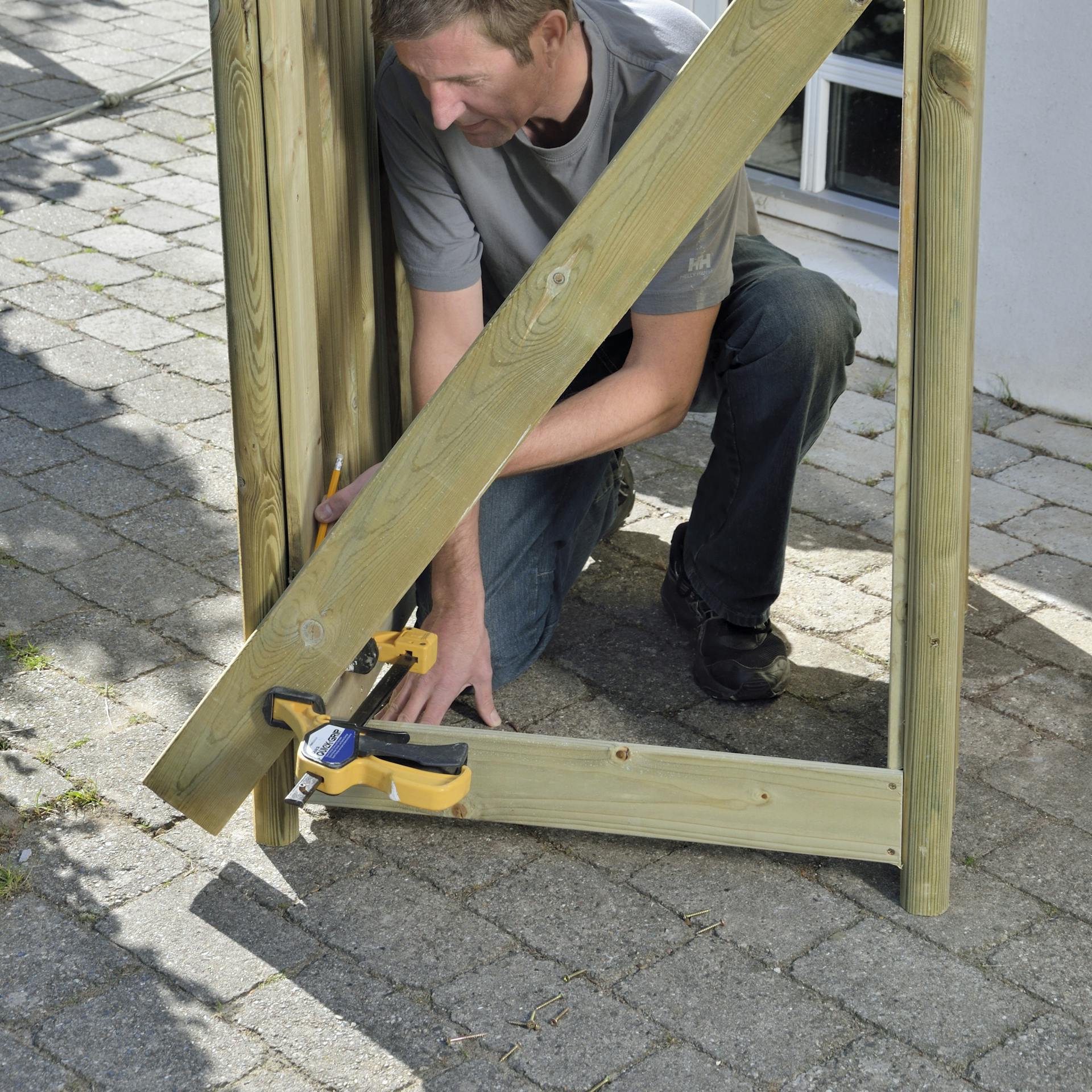 Osoba pripevňuje priečne laty k zadnej strane boxu na smetnú nádobu pomocou stolárskych svoriek