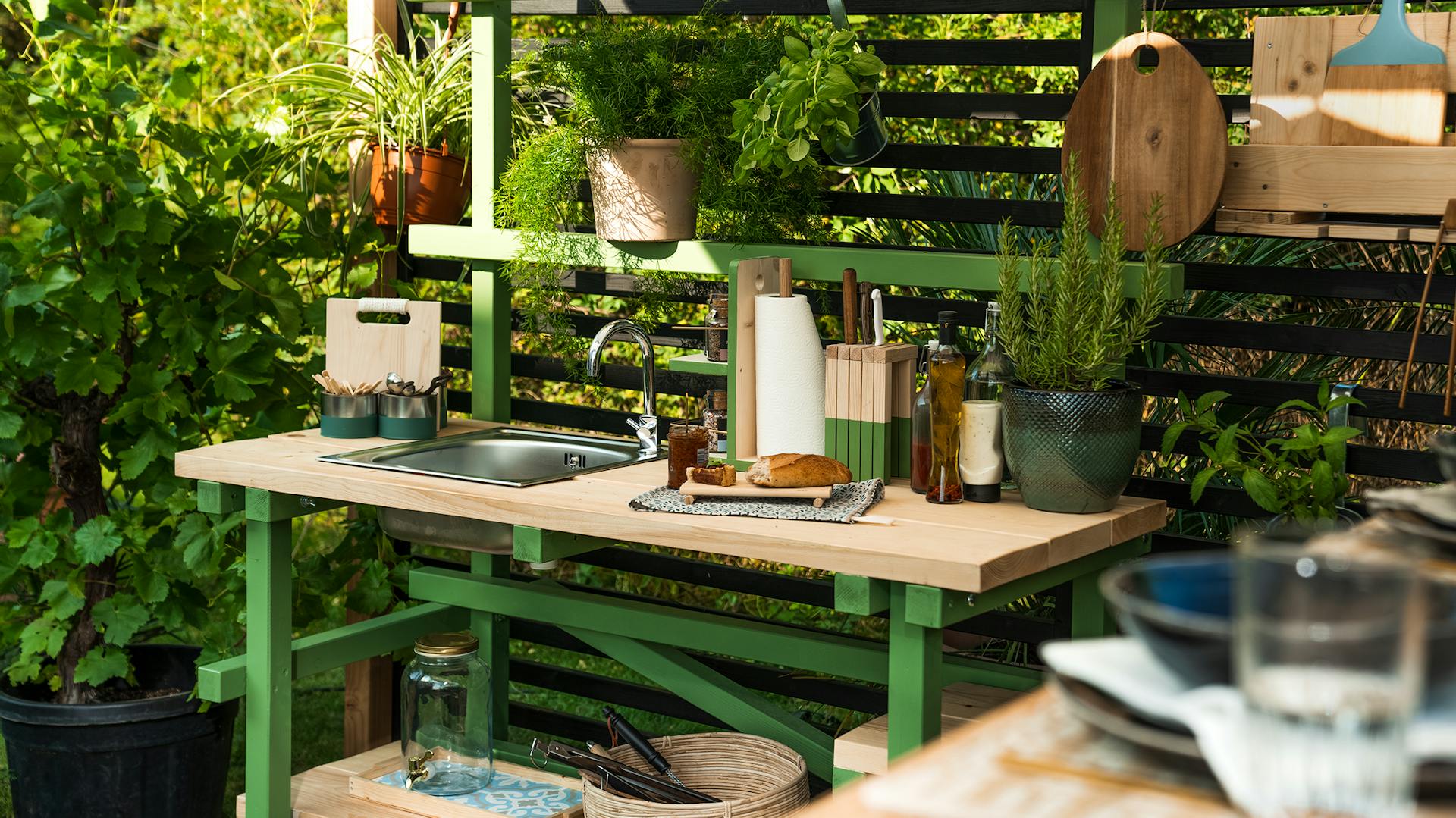 Outdoorküche Alfons, welche hellgrün gestrichen worden ist, steht in einer Ecke eines sommerlichen Gartens.