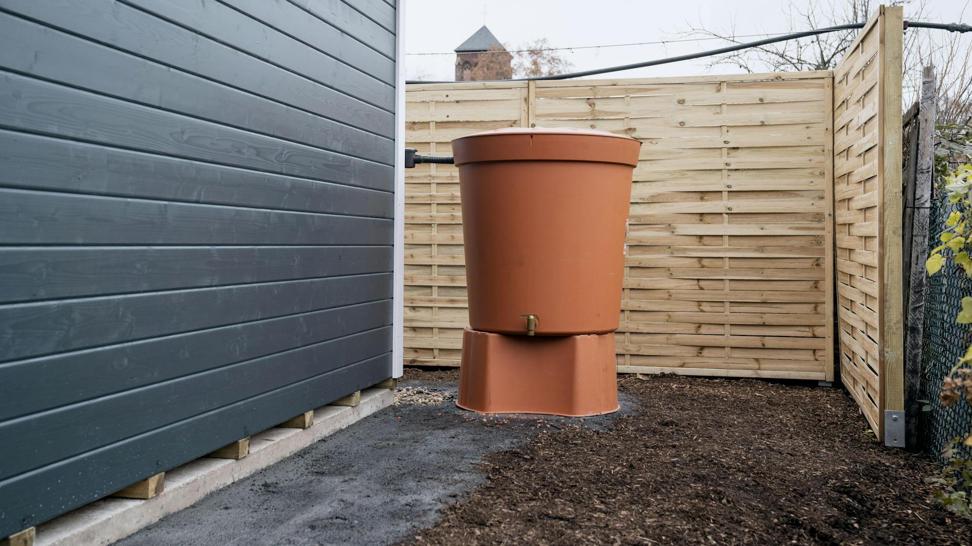 Eine angeschlossene und terrakottafarbende Regentonne steht in der Ecke vor einer holzvertäfelten und dunkelgrau gestrichenen Hauswand und einem holzfarbenden Palisadenzaun.