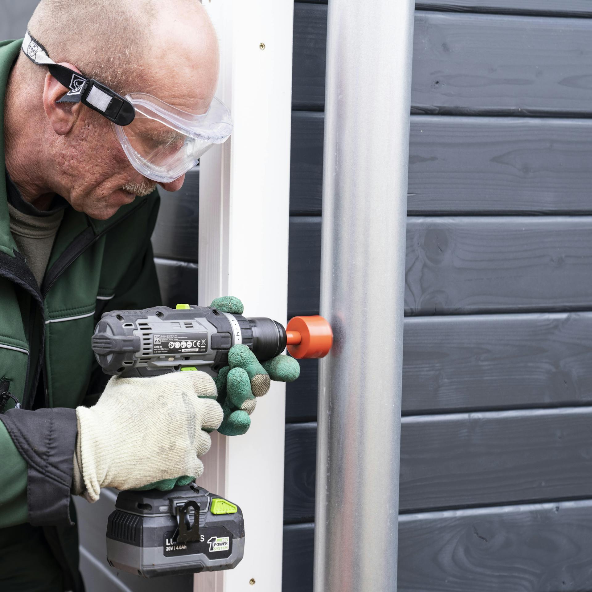 Handwerker mit Schutzbrille und Arbeitshandschuhen bohrt mit einem Kronenbohrer ein Loch in das Fallrohr.