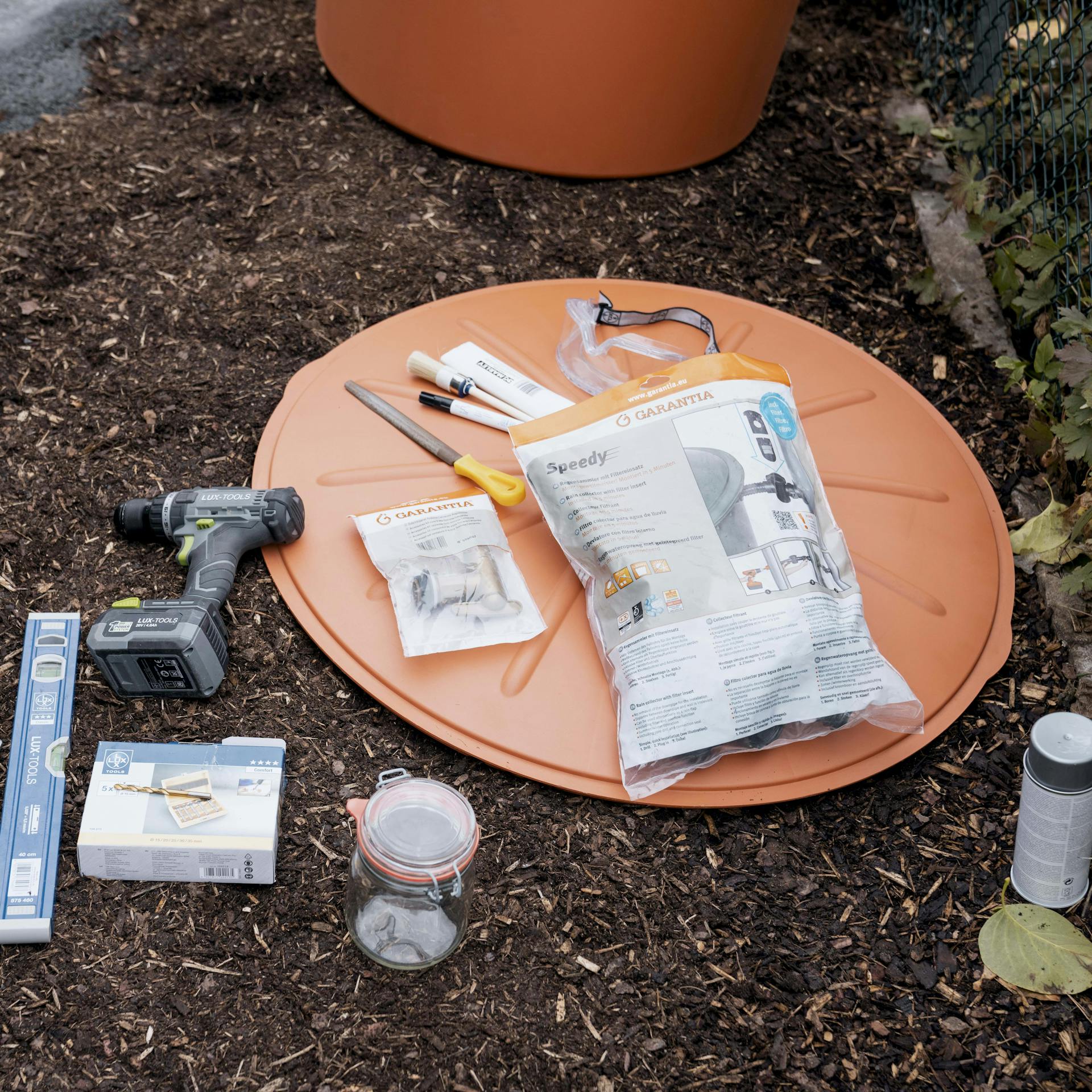 Alle benötigten Materialien zum Aufstellen einer Regentonne liegen ausgebreitet und griffbereit auf Rindenmulch.