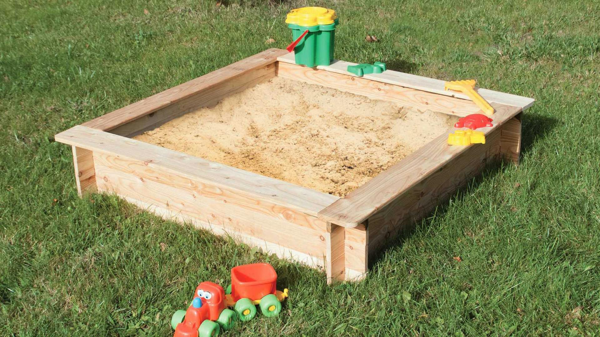Ein hölzerner Sandkasten mit Spielzeug steht auf einem Rasenstück.