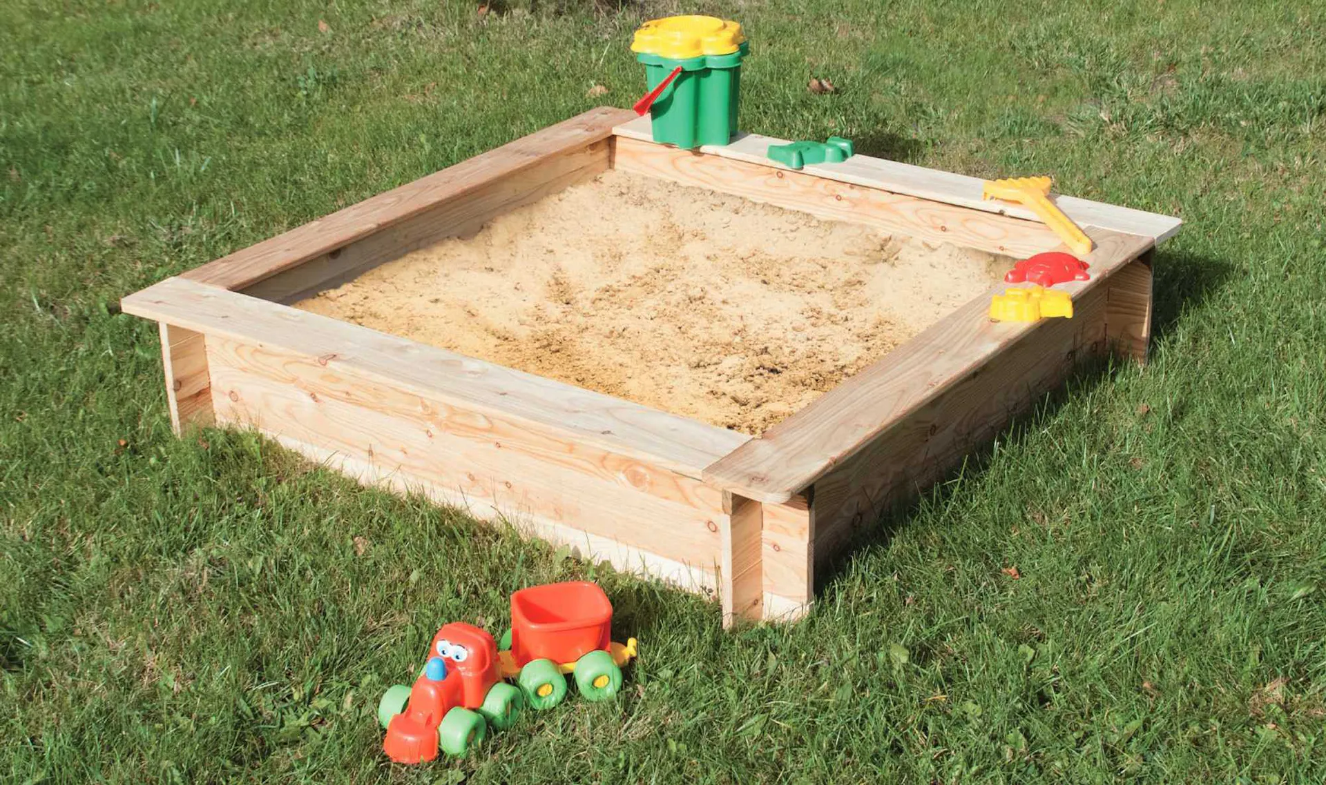 Sabbiera in legno riempita di sabbia e con giochi da spiaggia su prato verde.