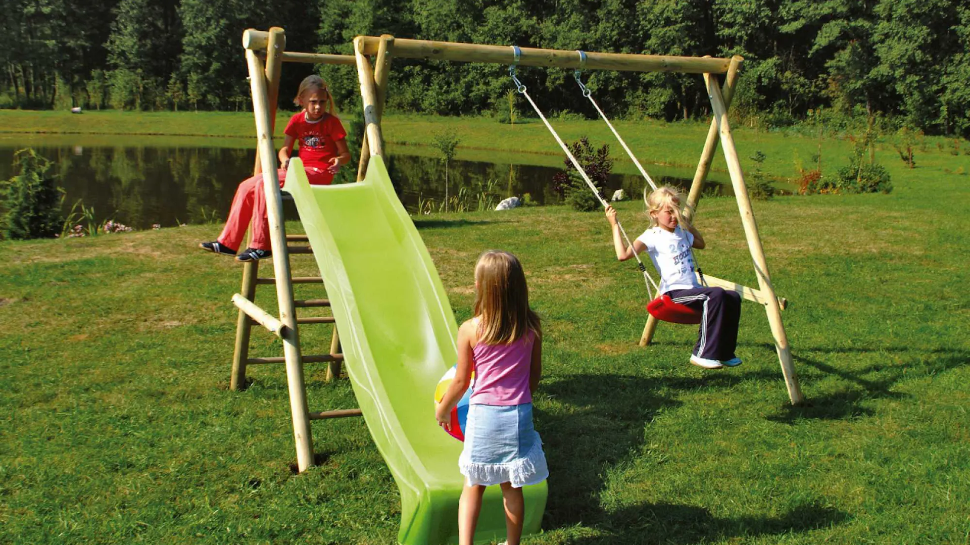 Tre bambini giocano su parco giochi in legno con un’altalena e uno scivolo verde in giardino.