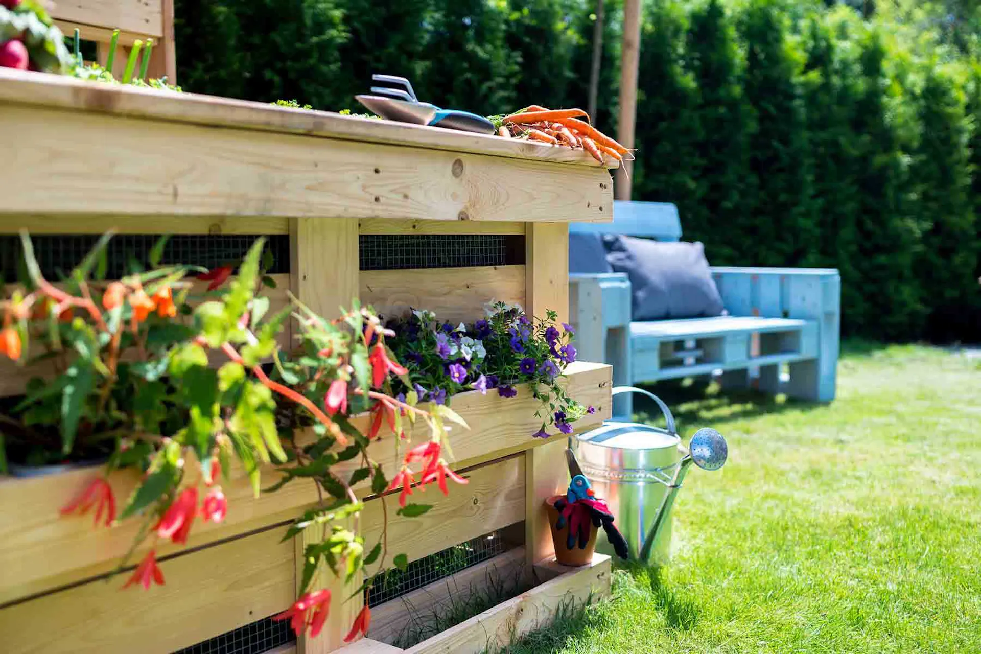 Hochbeet aus Paletten mit Pflanzen befüllt neben Giesskanne und Palettencouch in Garten