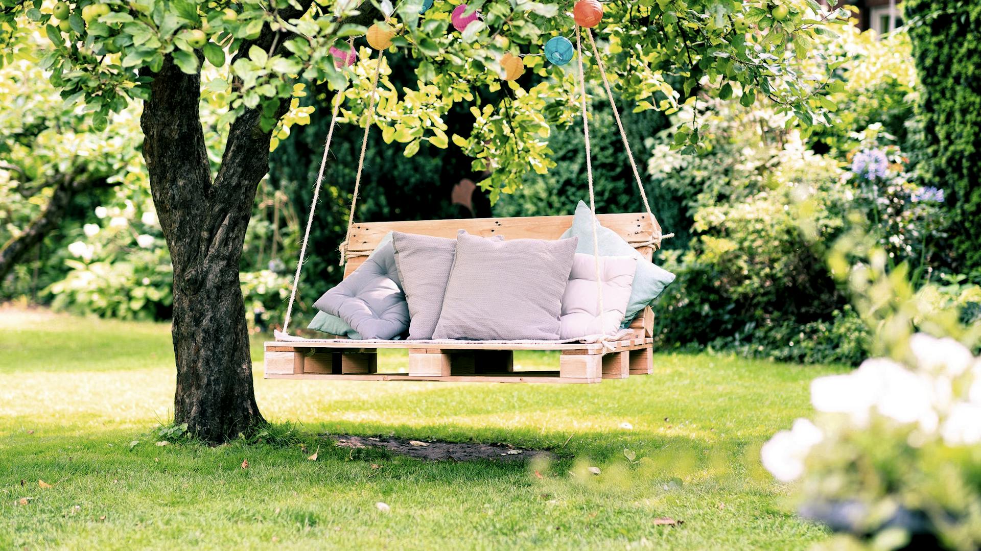 Hollywoodschaukel aus Paletten mit Kissen und Lampions geschmückt an Baum hängend im Garten