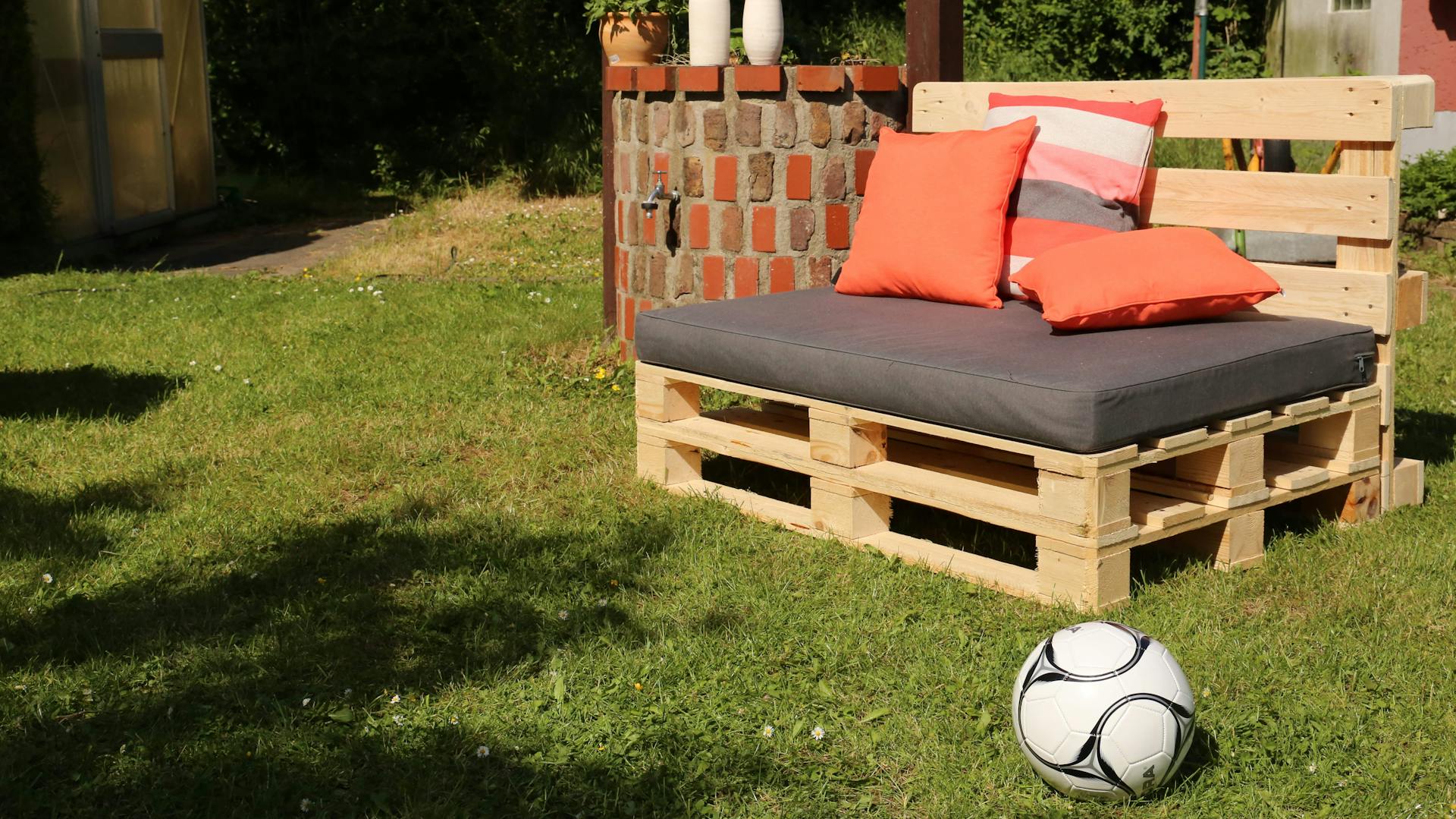 Paletten-Sofa mit grauen und orangenen Kissen in Garten neben Fußball