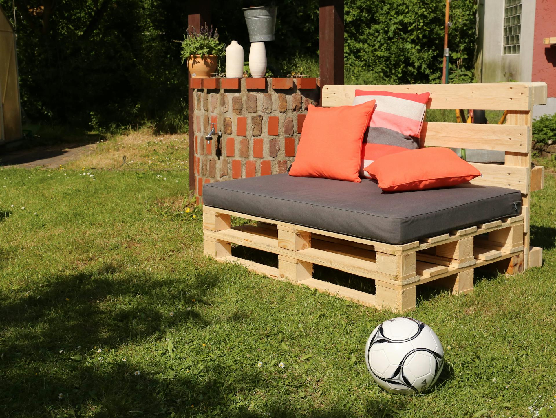 Divano in pallet con cuscini grigi e arancioni in giardino accanto al pallone da calcio