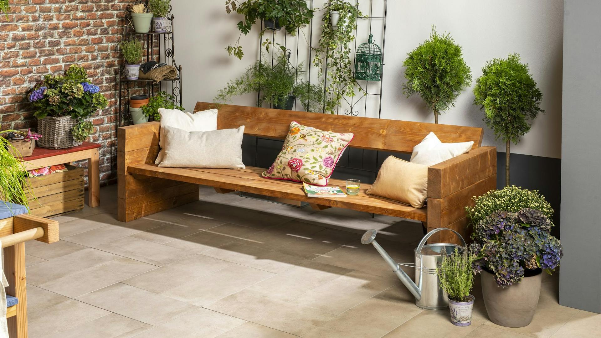 Selbstgebaute Bank aus Teakholz mit drapierten Sitzkissen auf Terrasse umringt von Pflanzen