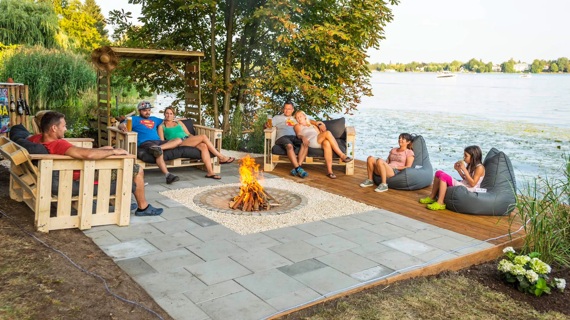 Personen sitzen auf Gartenmöbeln rund um Feuerstelle im Garten am See.