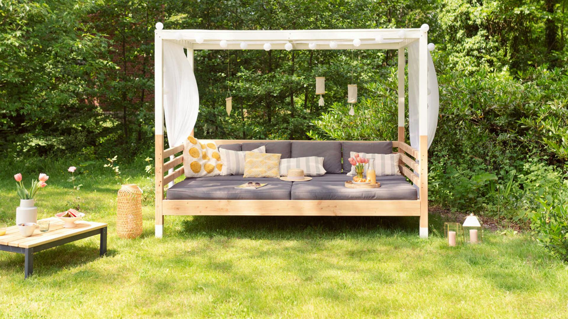 Frontansicht der aus Vollholz selbstgebauten Sonneninsel Helena in naturfarbe und mit weißem Baldachin mit Sitzpolstern.