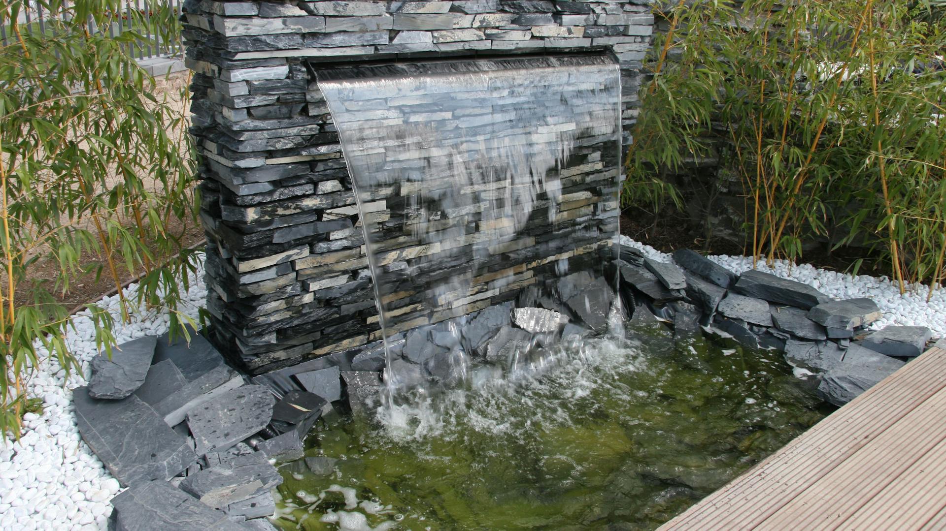 Moderner, frei fallender Wasserfall fließt aus Schiefermauer in Wasserbecken.
