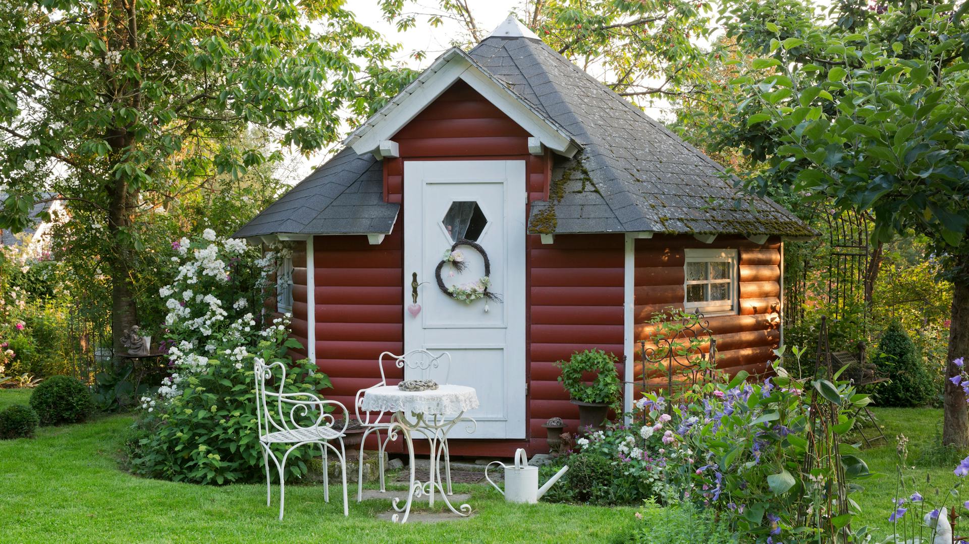 Ein rotes Gartenhaus in einem Garten mit weißen Gartenstühlen und einem Tisch