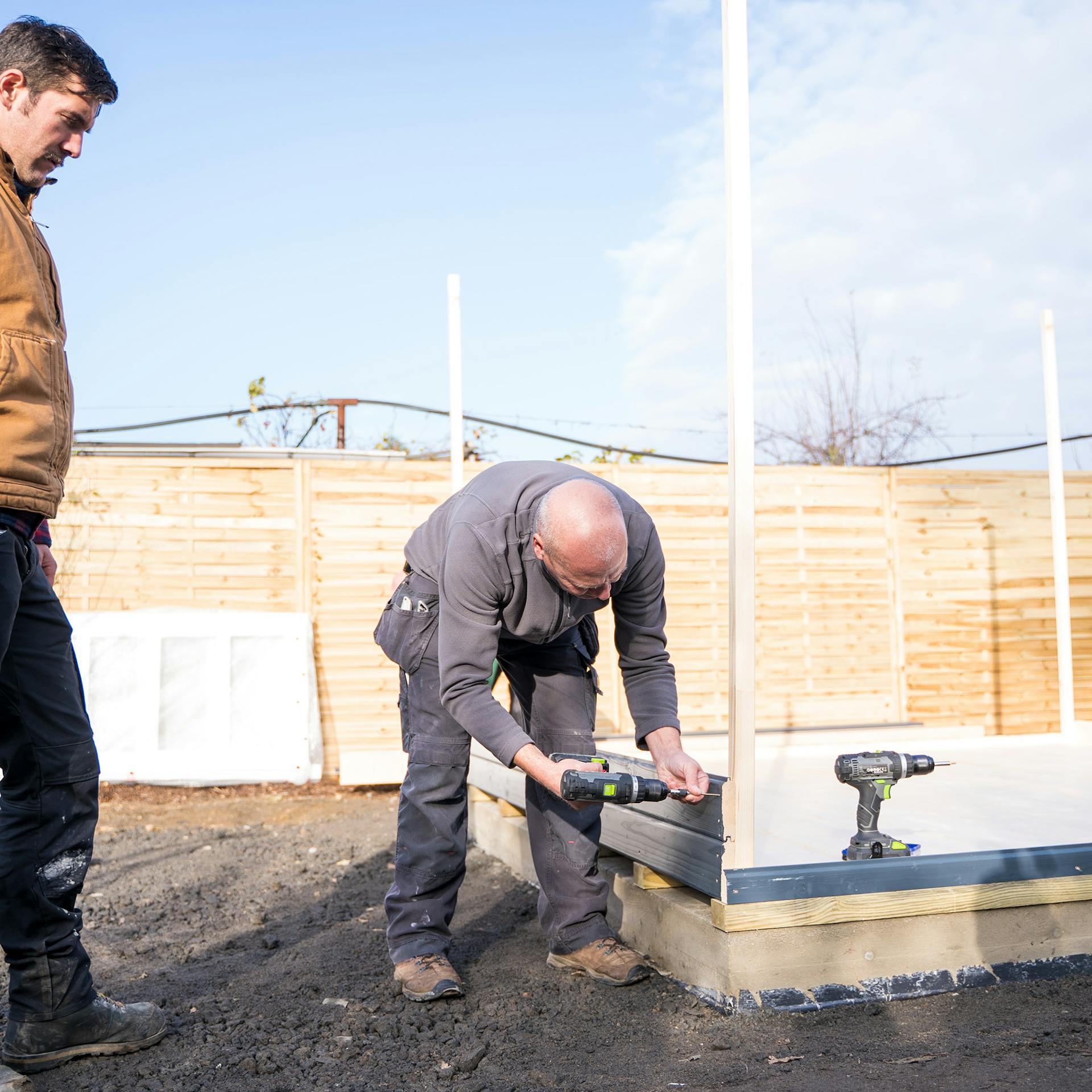Egy ember csaphornyos deszkákkal szereli össze a kerti ház oldalfalait, és a sarokoszlopokhoz rögzíti őket.