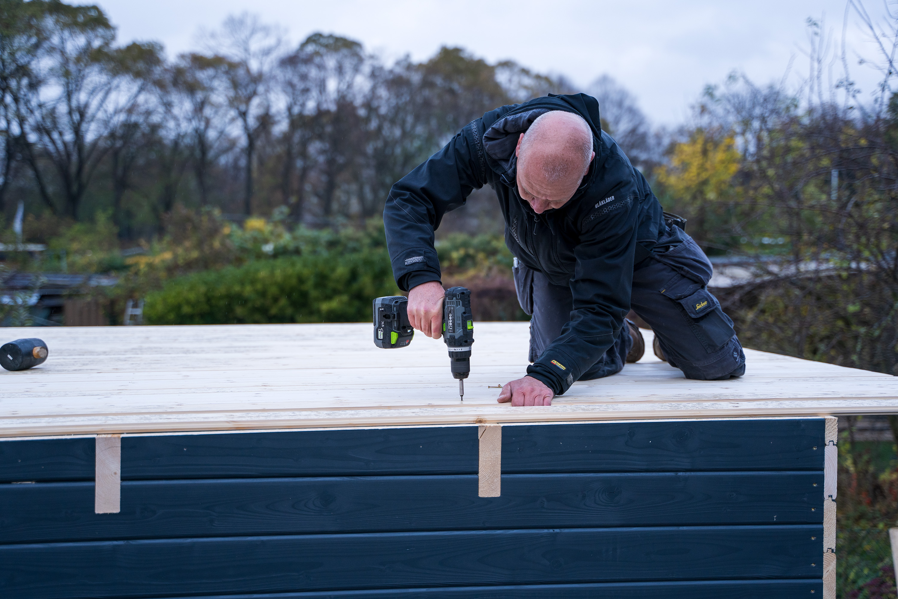 Egy ember tetődeszkákat csavaroz fel a tetőgerendákra egy akkus csavarozóval.