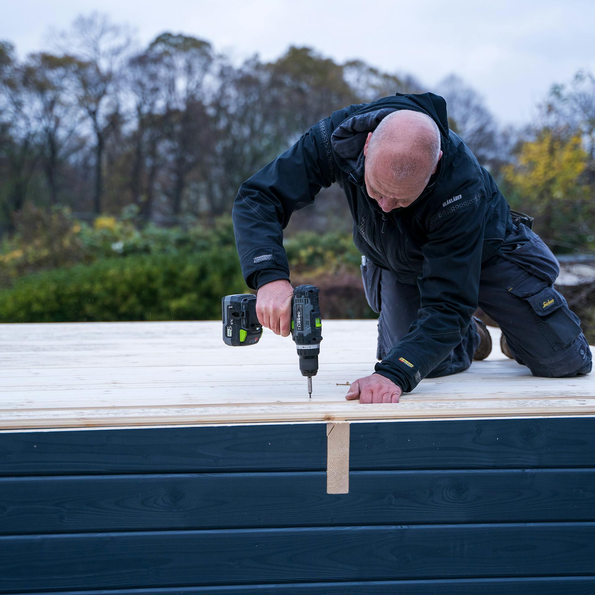 Egy ember tetődeszkákat csavaroz fel a tetőgerendákra egy akkus csavarozóval.