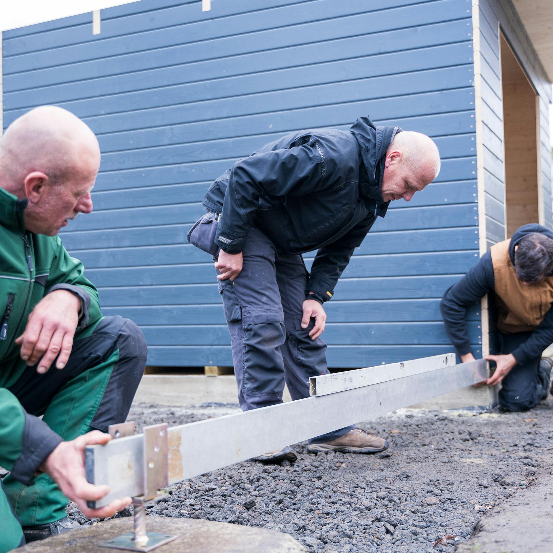 Három ember beállítja a sarokoszlopot és vízmértékkel ellenőrzi azt.