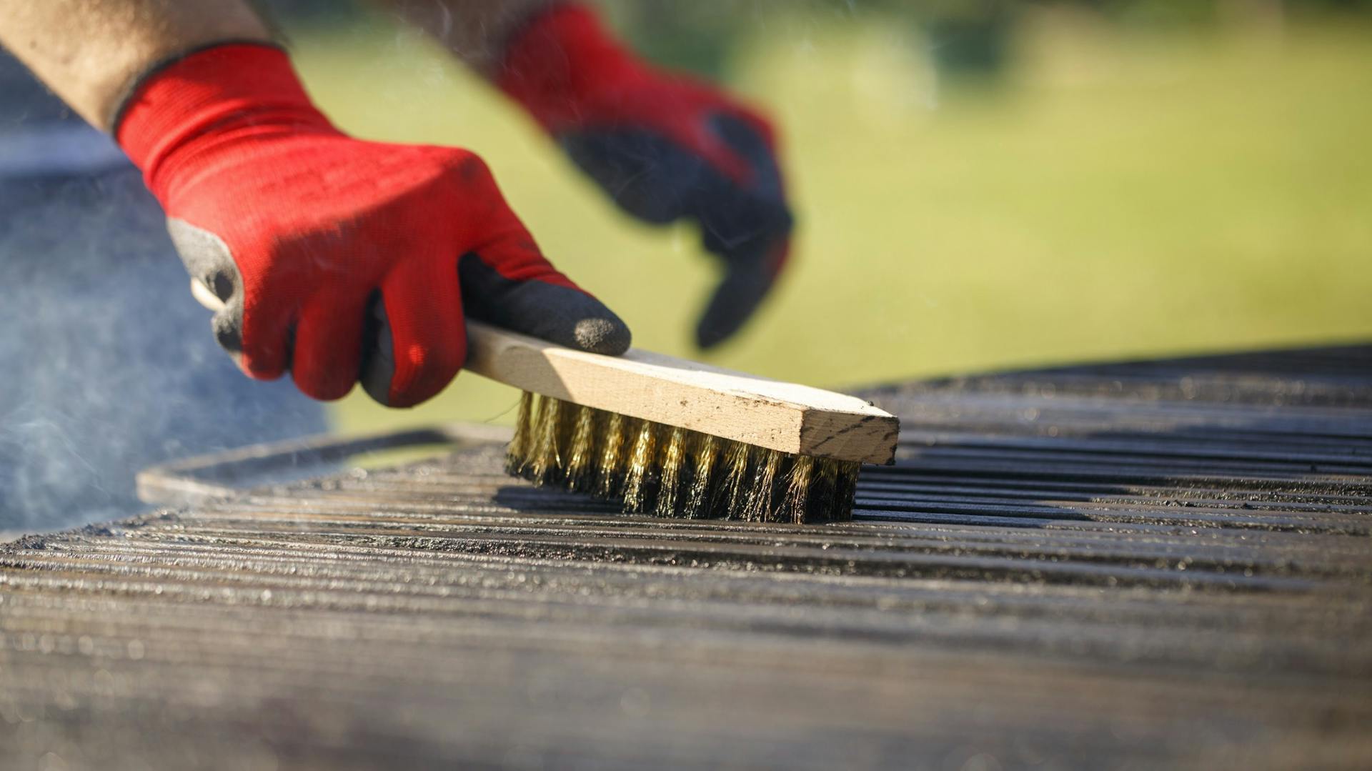 Mann trägt rote Schutzhandschuhe und reinigt Grill mit Drahtbürste.