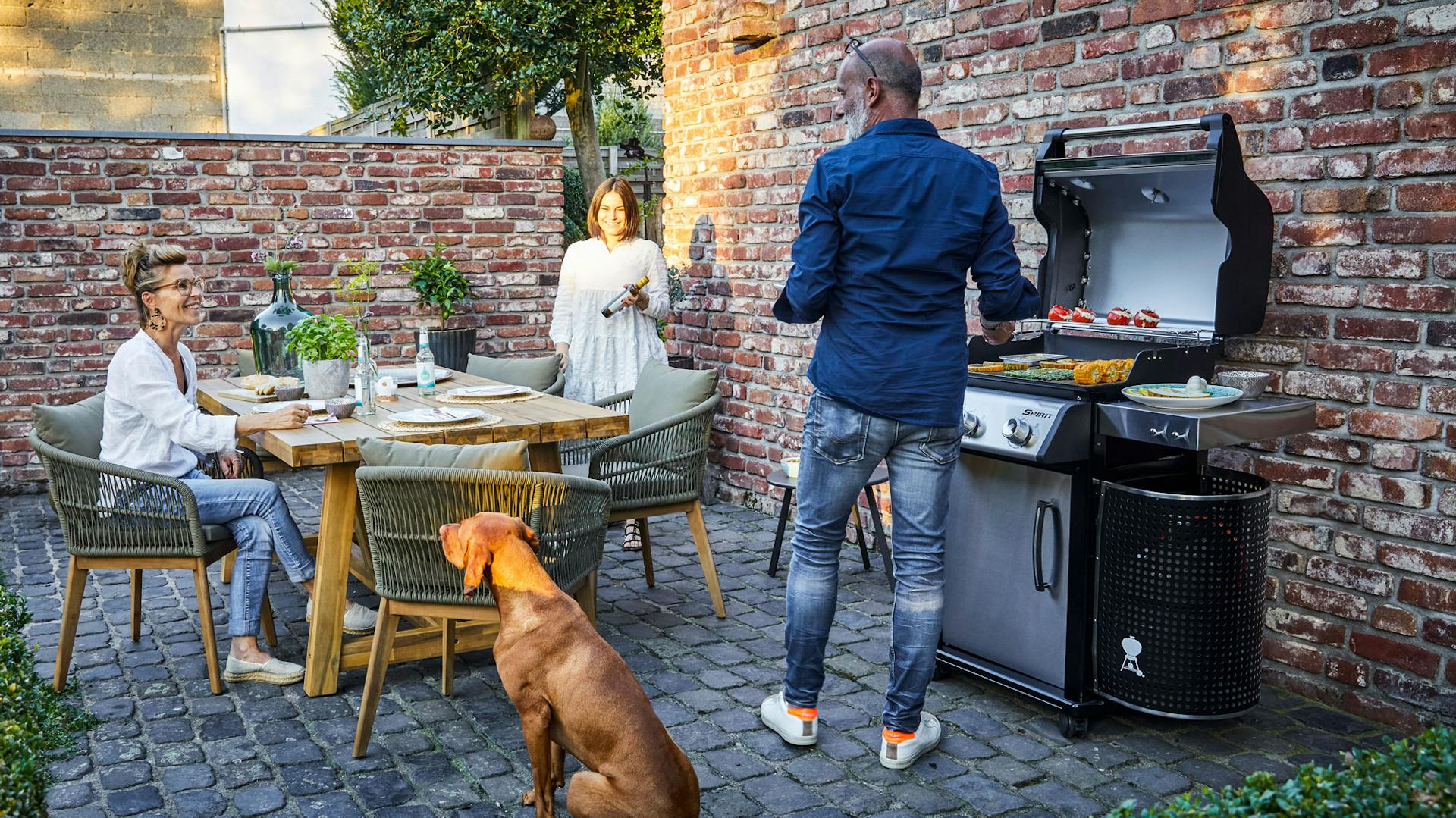 Mann grillt am Gasgrill auf Terrasse mit Hund