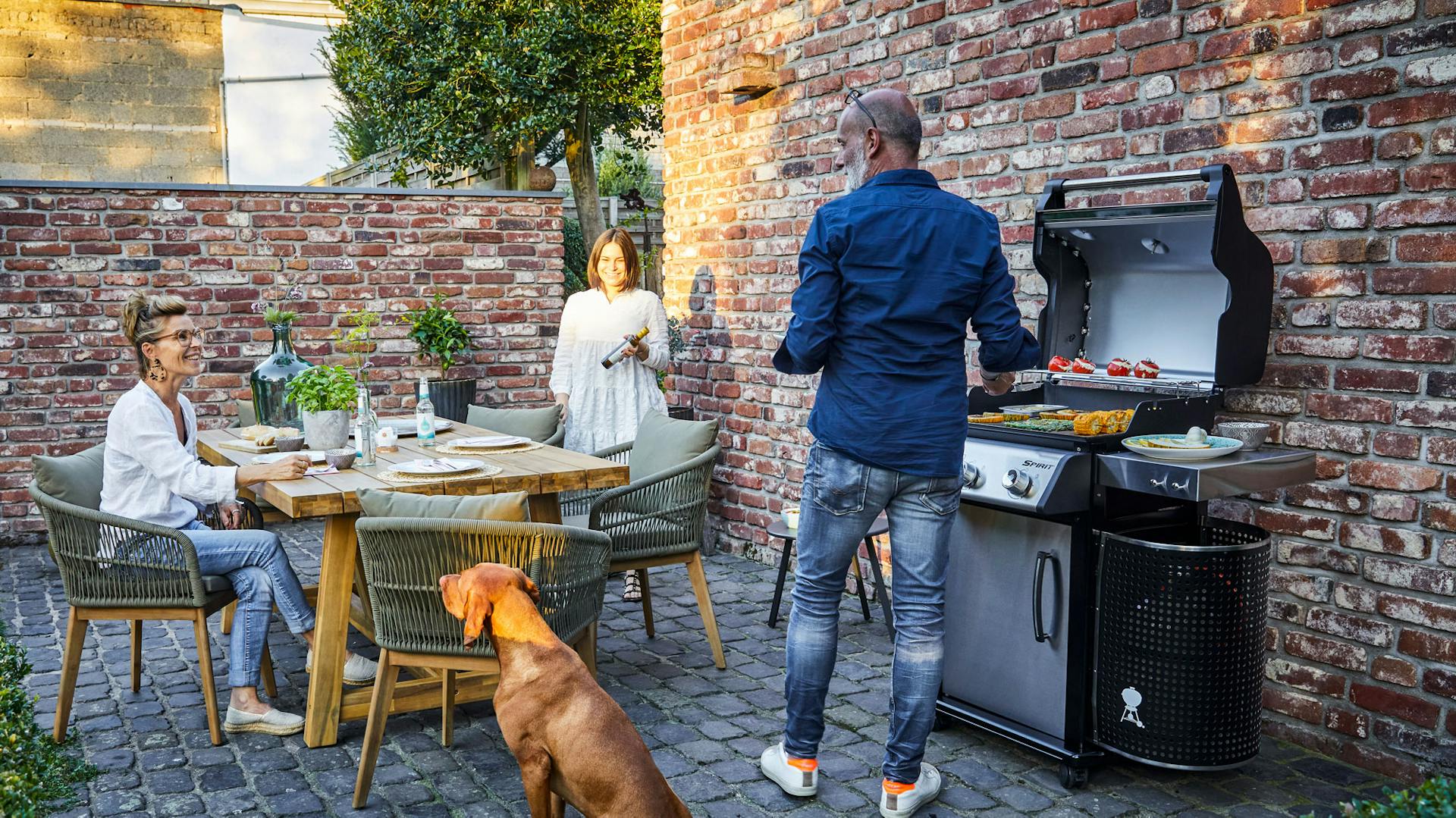 Mann grillt am Gasgrill auf Terrasse mit Hund