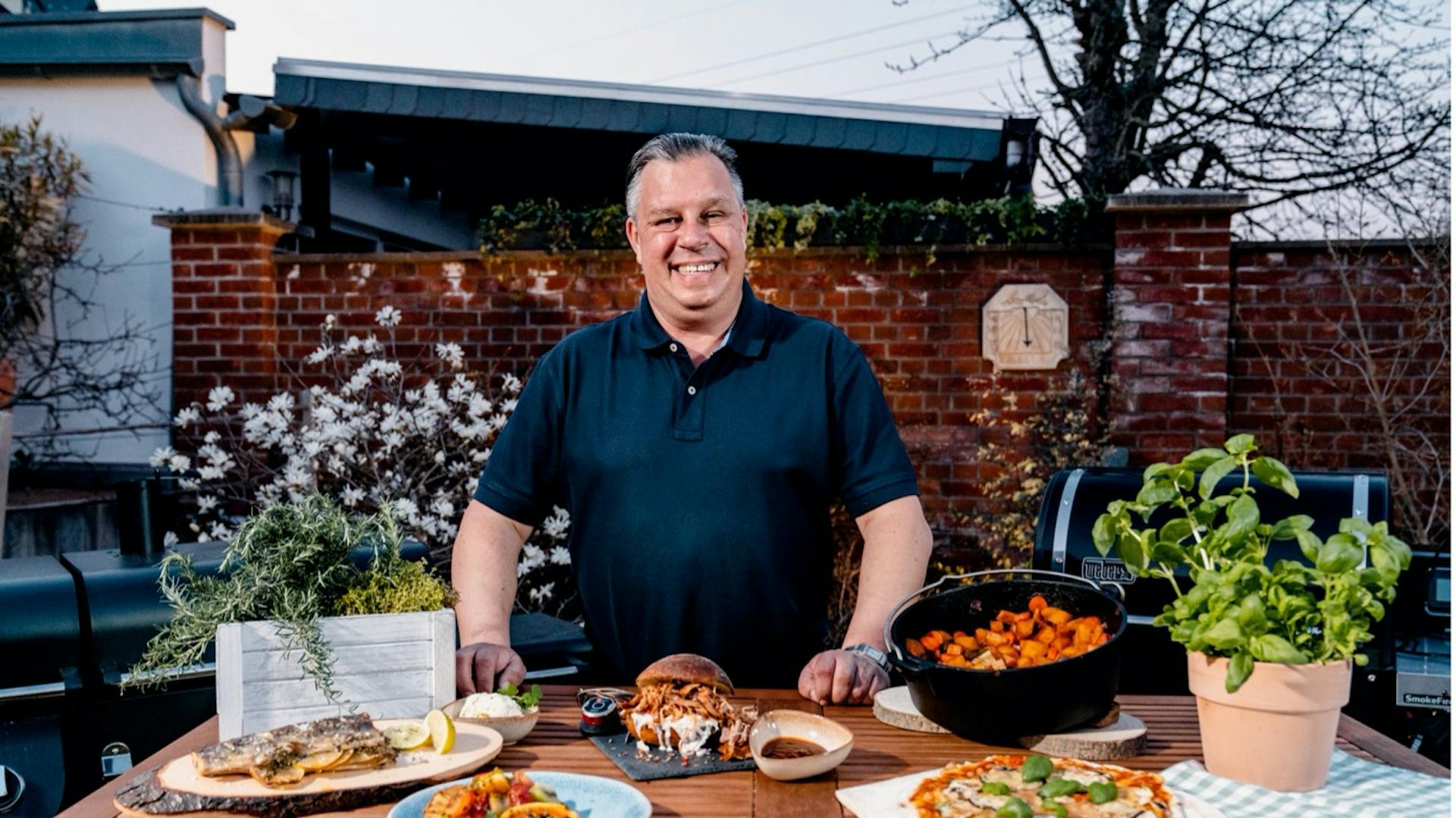 Grillmeister Michael Hoffmann an Gartentisch mit gegrillten Speisen und Zutaten.