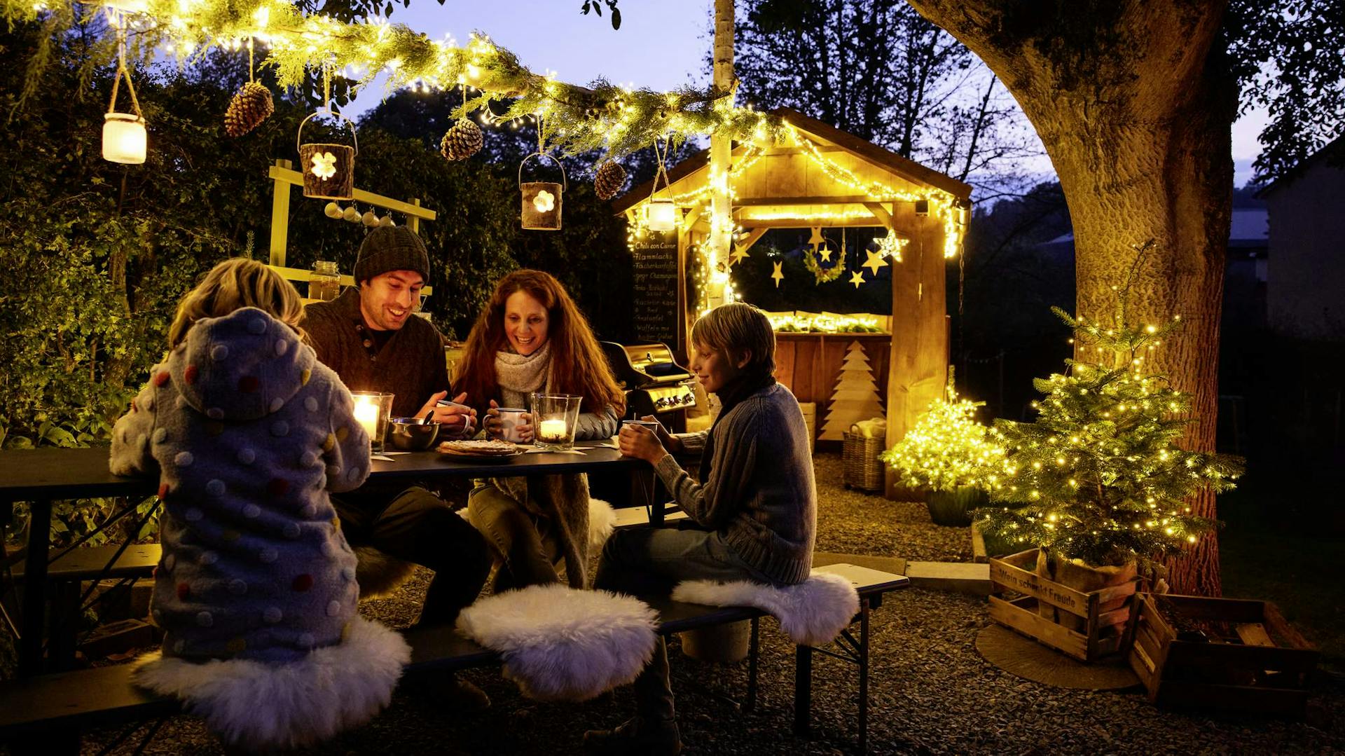 Personen sitzen auf einer Bierbankgarnitur, der Garten ist winterlich dekoriert. In einer Ecke steht ein Weihnachtsbaum