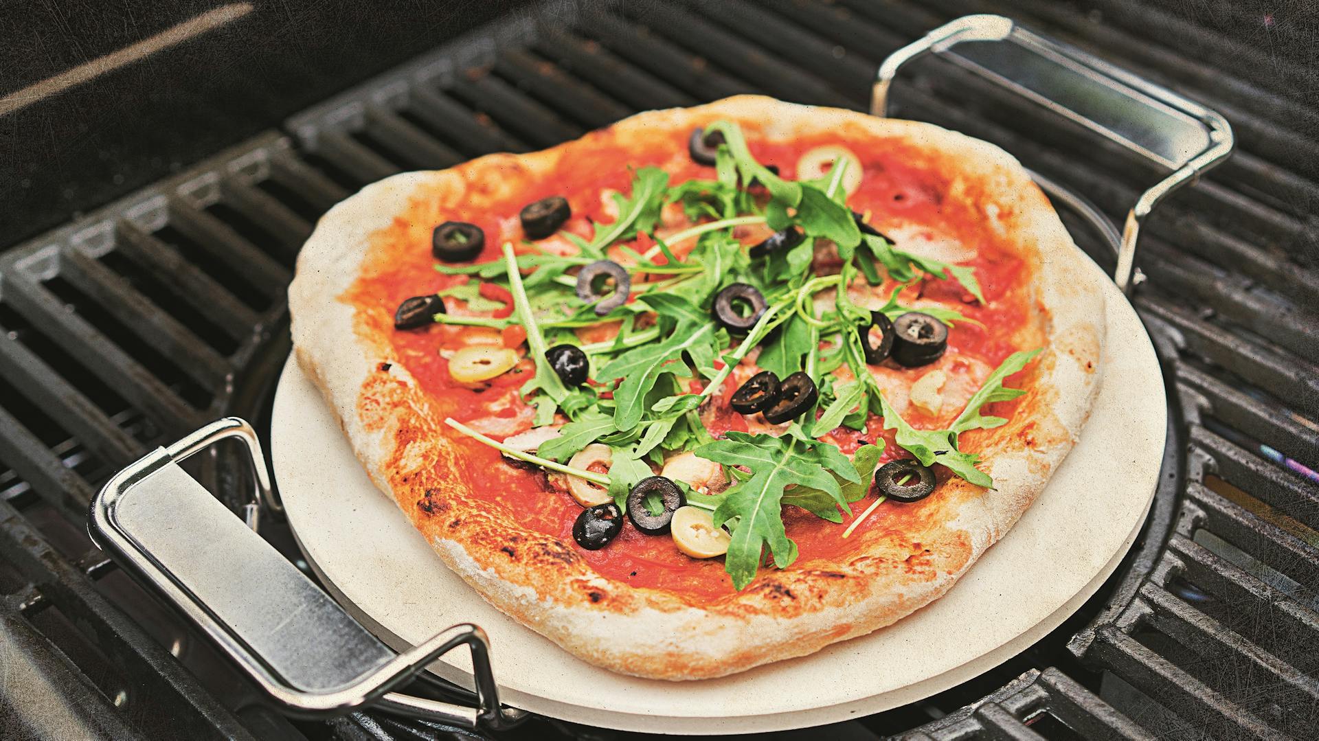 Belegte Pizza auf Pizzastein mit Henkeln auf Grill