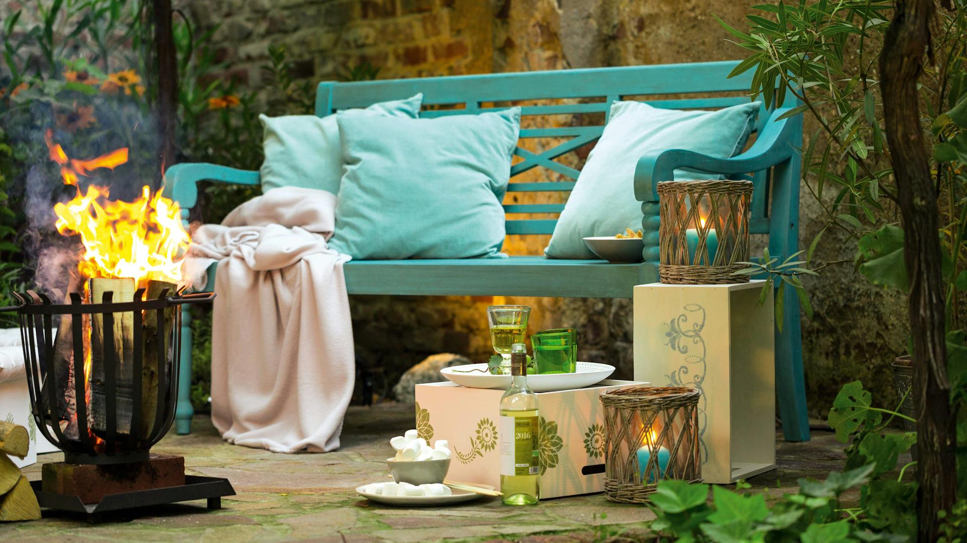 Gemütliche Sitzecke im Garten mit Bank, Kissen, Decke, Laternen und Feuerkorb