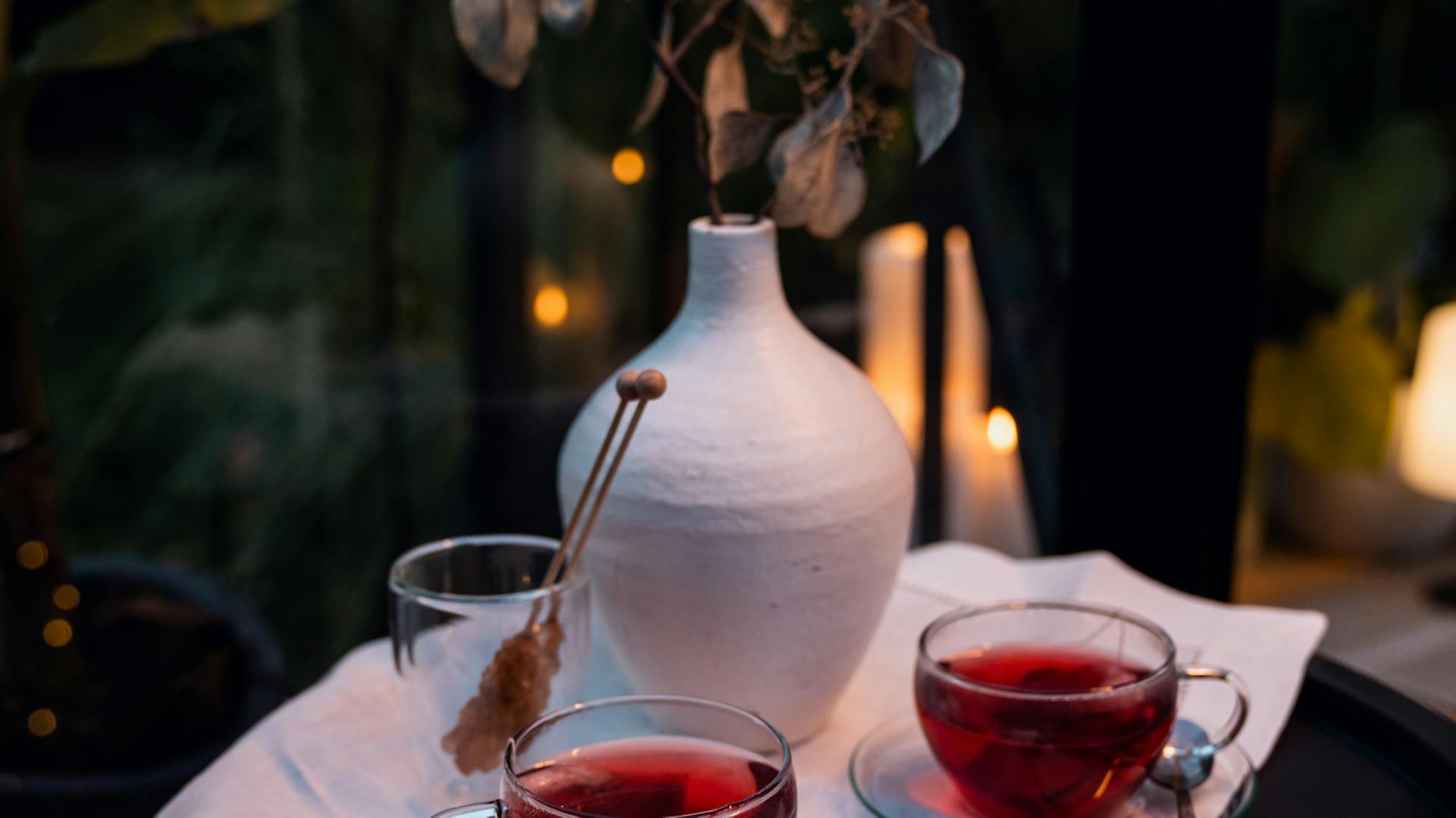 Zwei stimmungsvoll beleuchtete Tassen Tee stehen neben einer Vase vor dunklem Hintergrund.