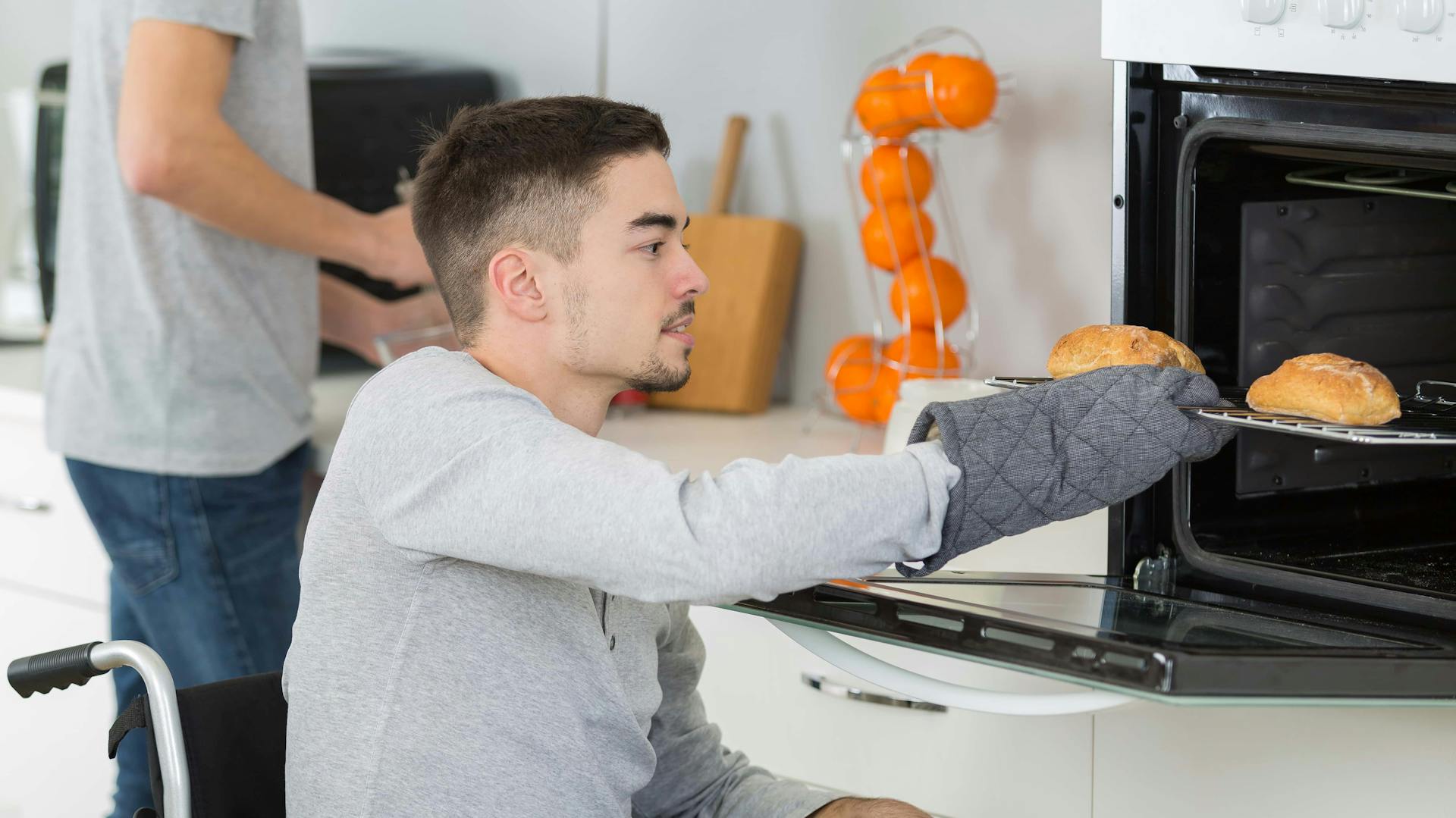 Ein Mann im Rollstuhl nimmt Essen aus einem höher positionierten Backofen heraus.