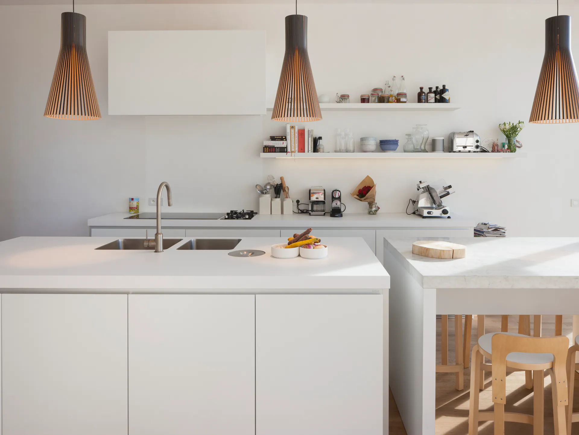 Küchenzeile mit weißen Fronten und weißer Arbeitsplatte. Rechts ein Tisch mit Holzstühlen. Drei Lampen hängen von der Decke.